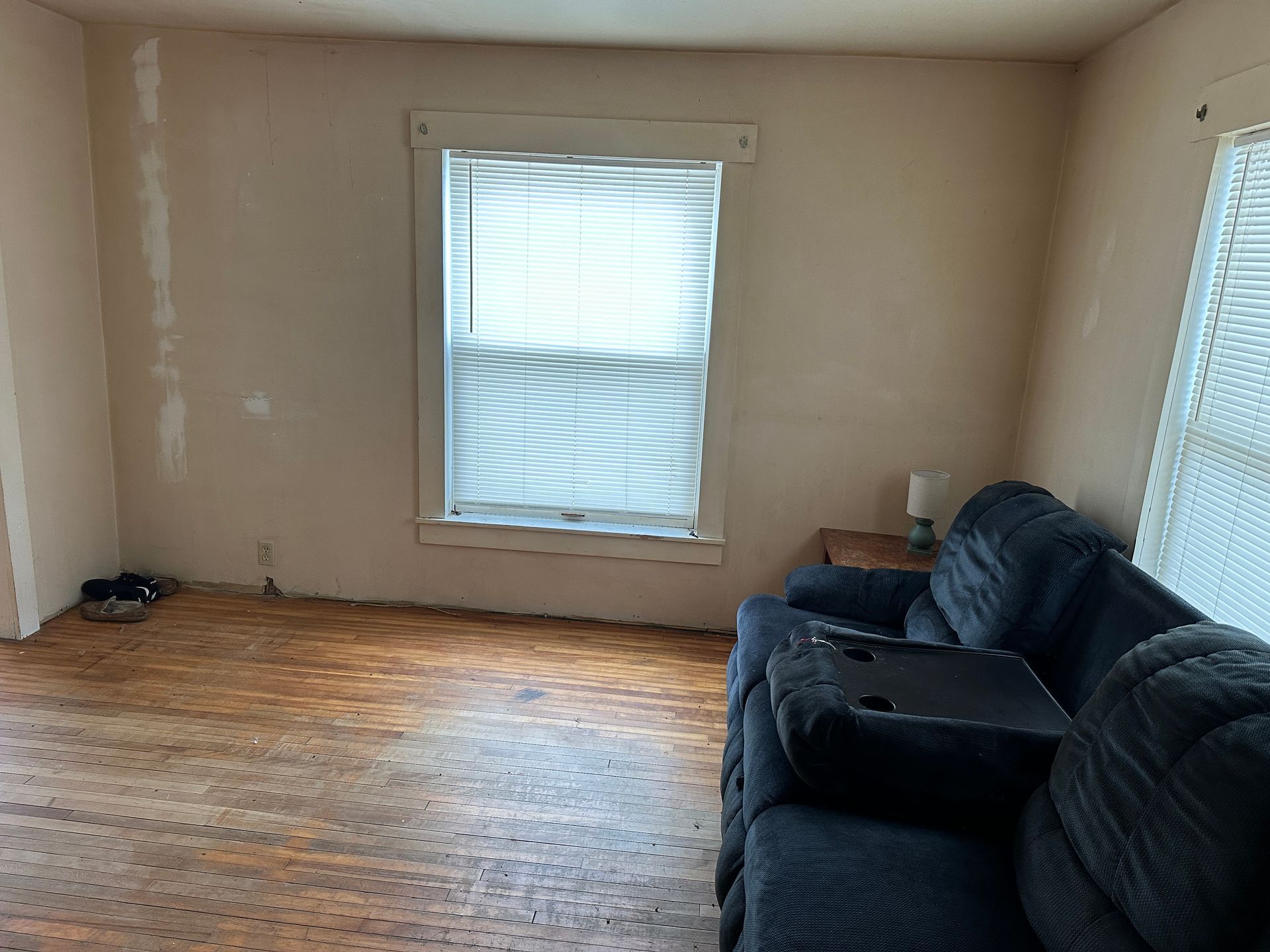 A room with light beige walls, hardwood flooring, a window with closed white blinds, and a dark blue reclining sofa.