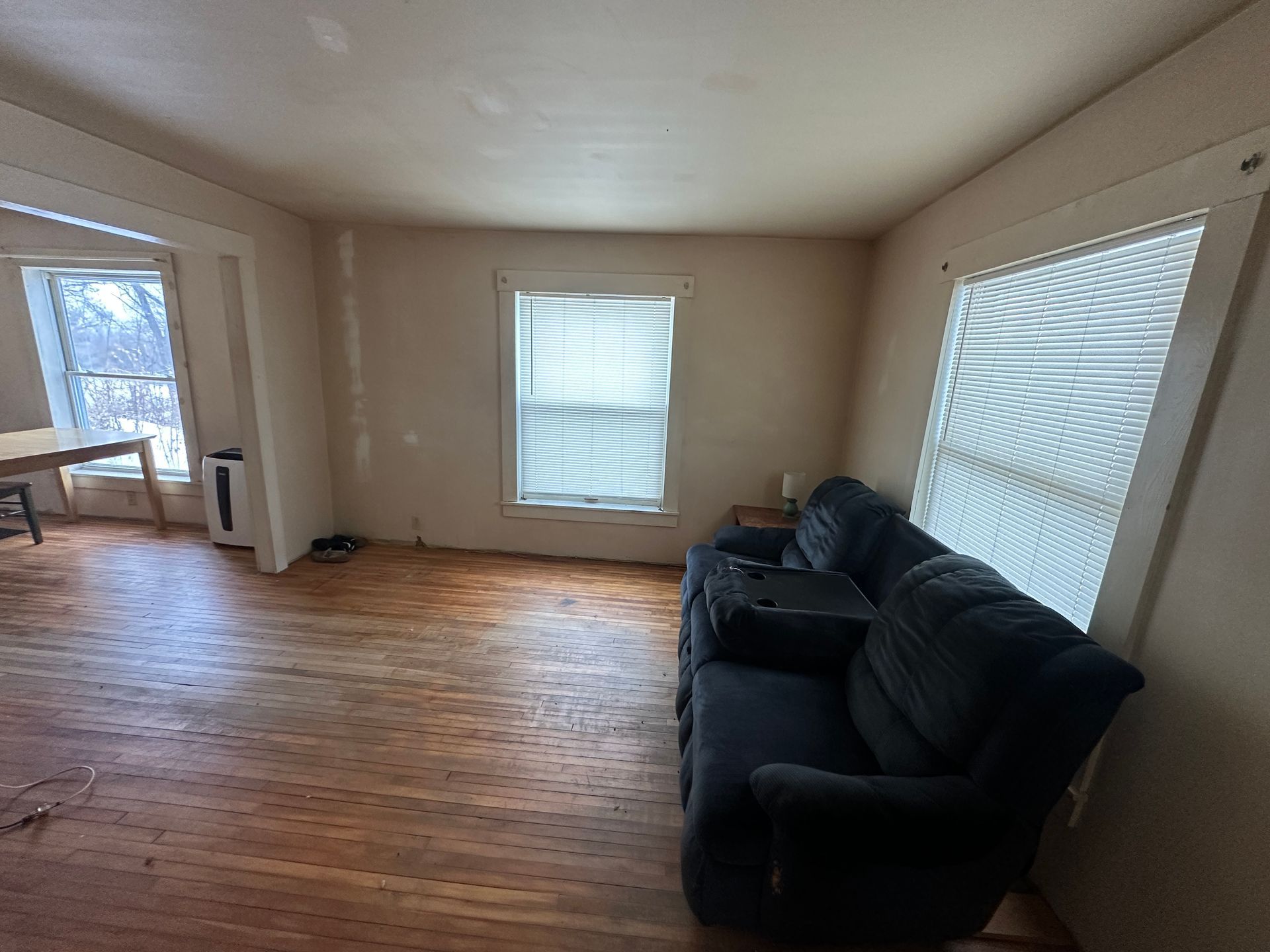 A living room with hardwood floors, a dark blue sofa against a wall, and windows with white blinds.