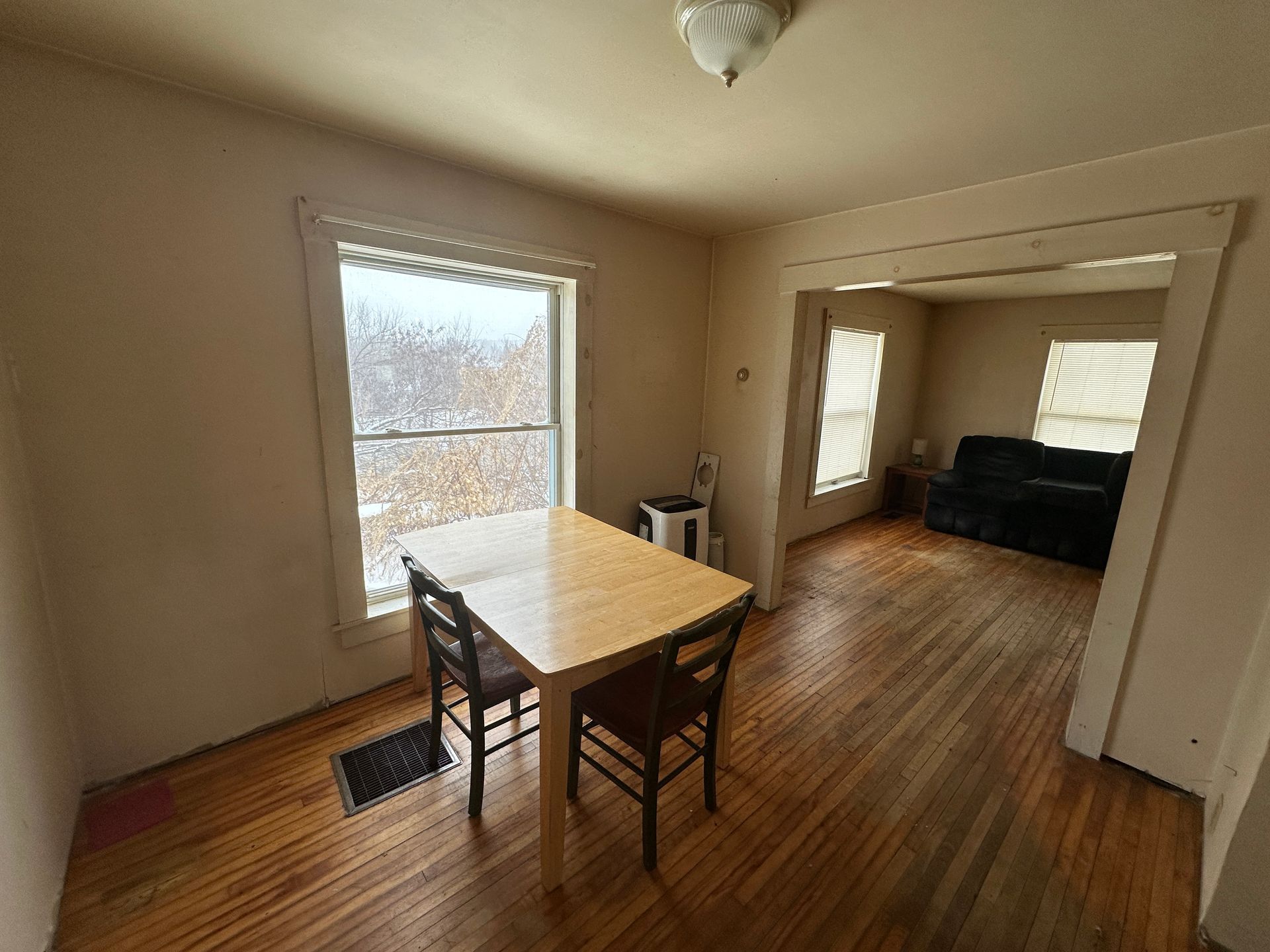 A dining room with a light wood table and two black chairs, opening into a living room with a dark couch on hardwood floors.