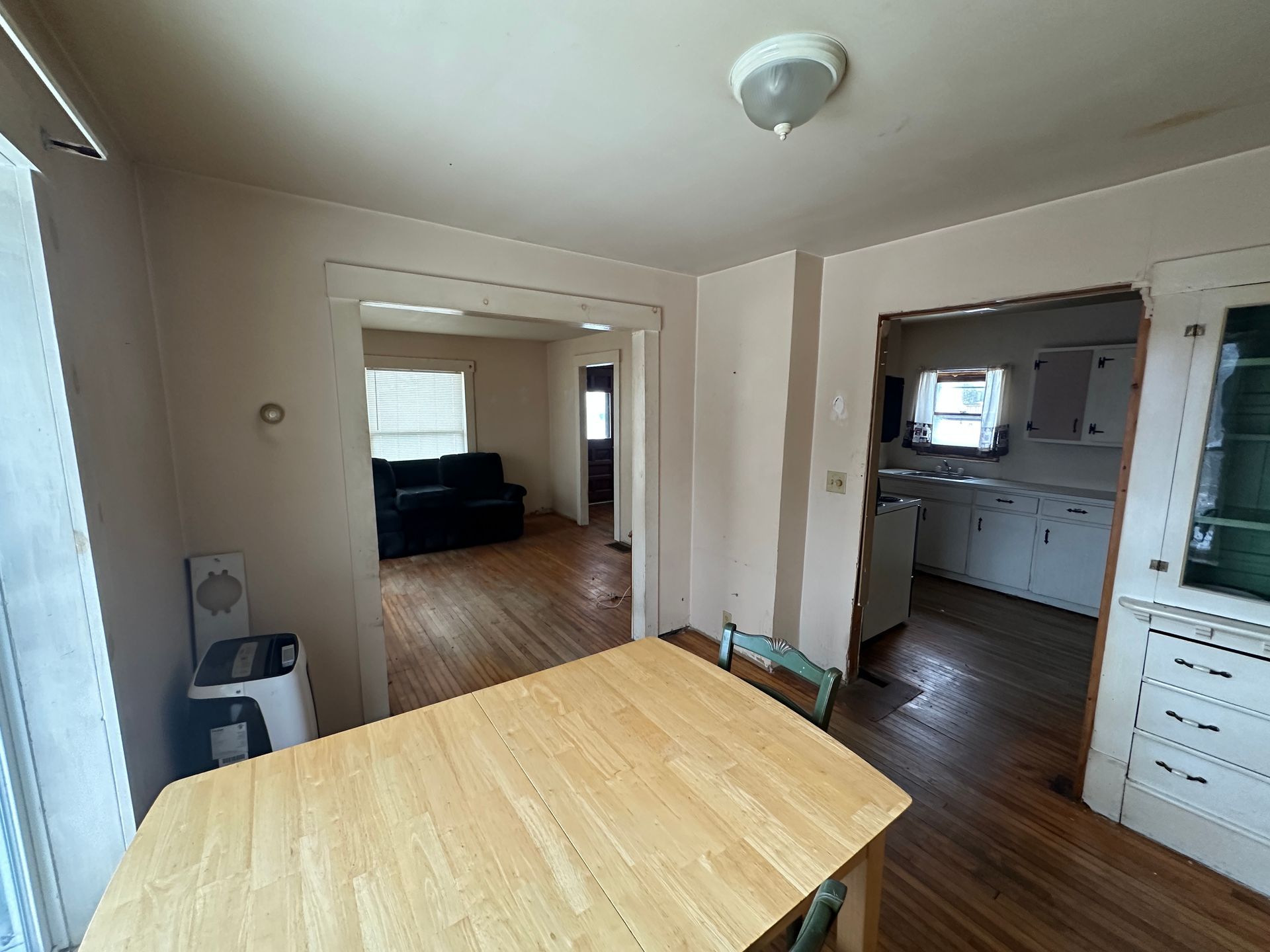 An interior view of a dining room with a light wood table, looking through doorways into a living room and a kitchen.