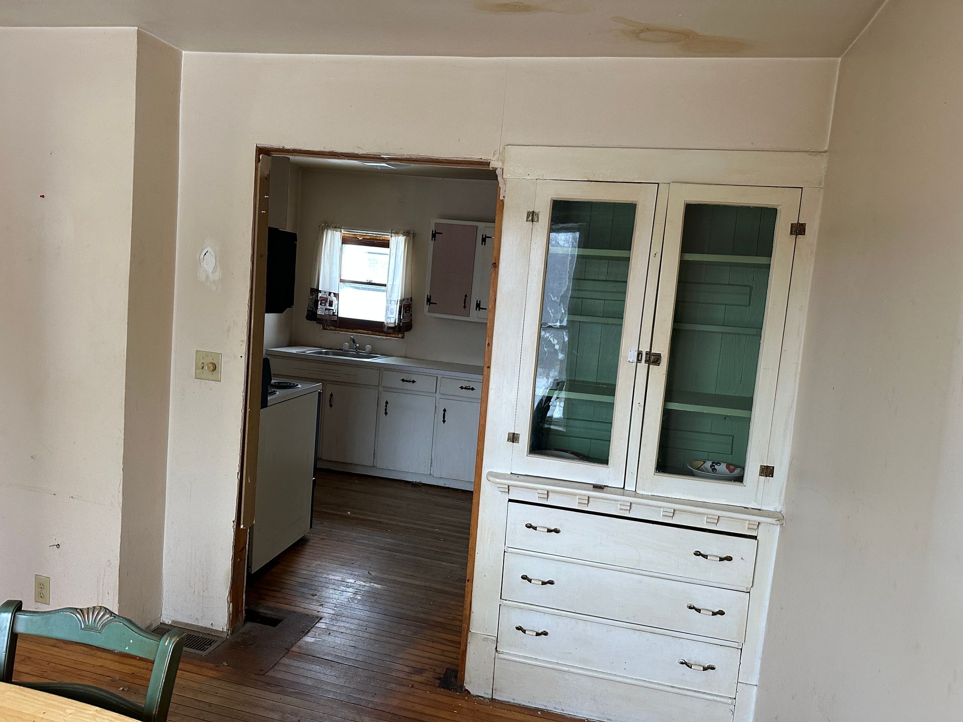 A white built-in hutch stands beside a doorway leading into a kitchen with white cabinetry and a window.