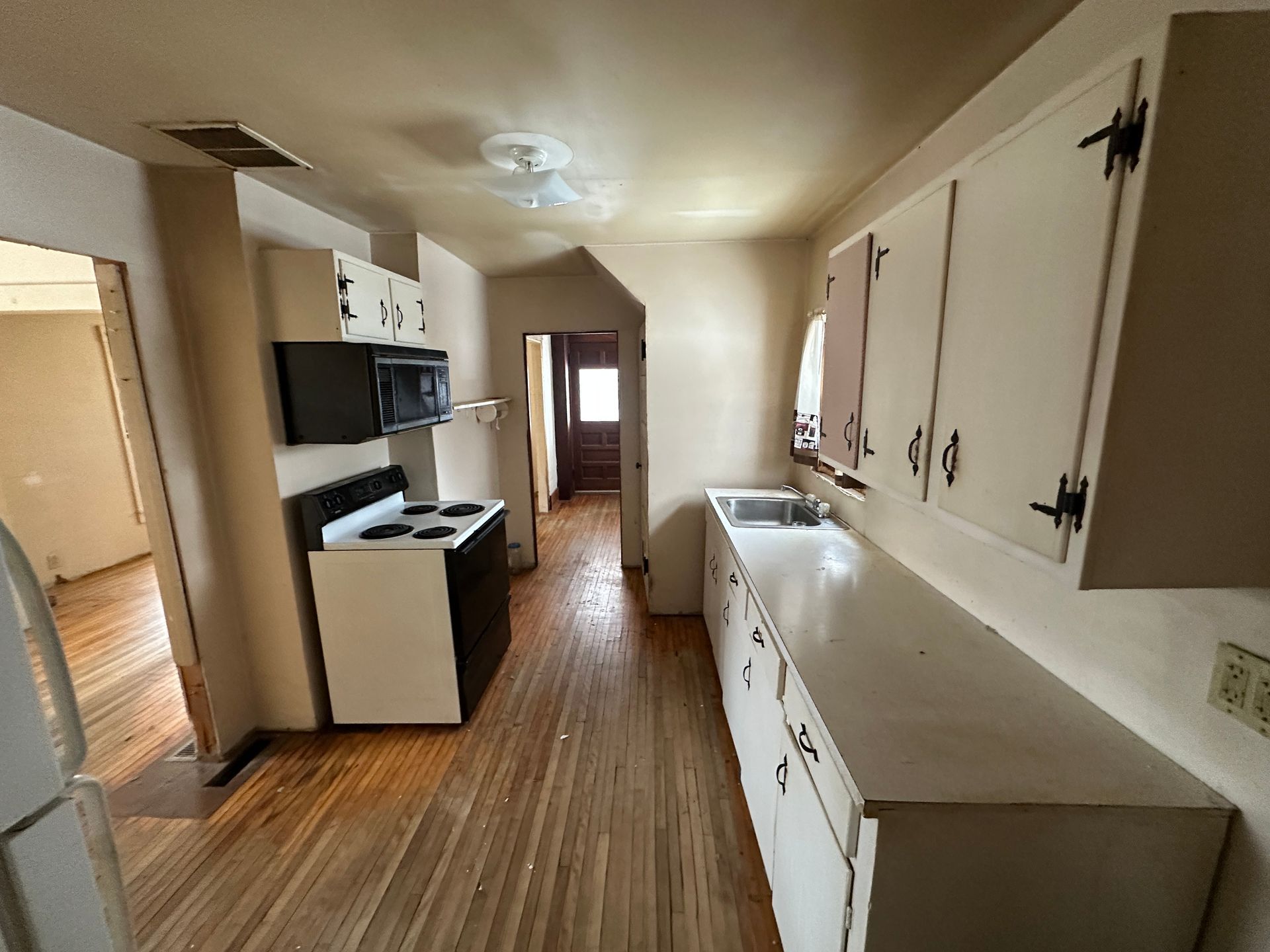 A long, narrow kitchen with white cabinets, a black and white stove, and hardwood floors leading to a doorway.