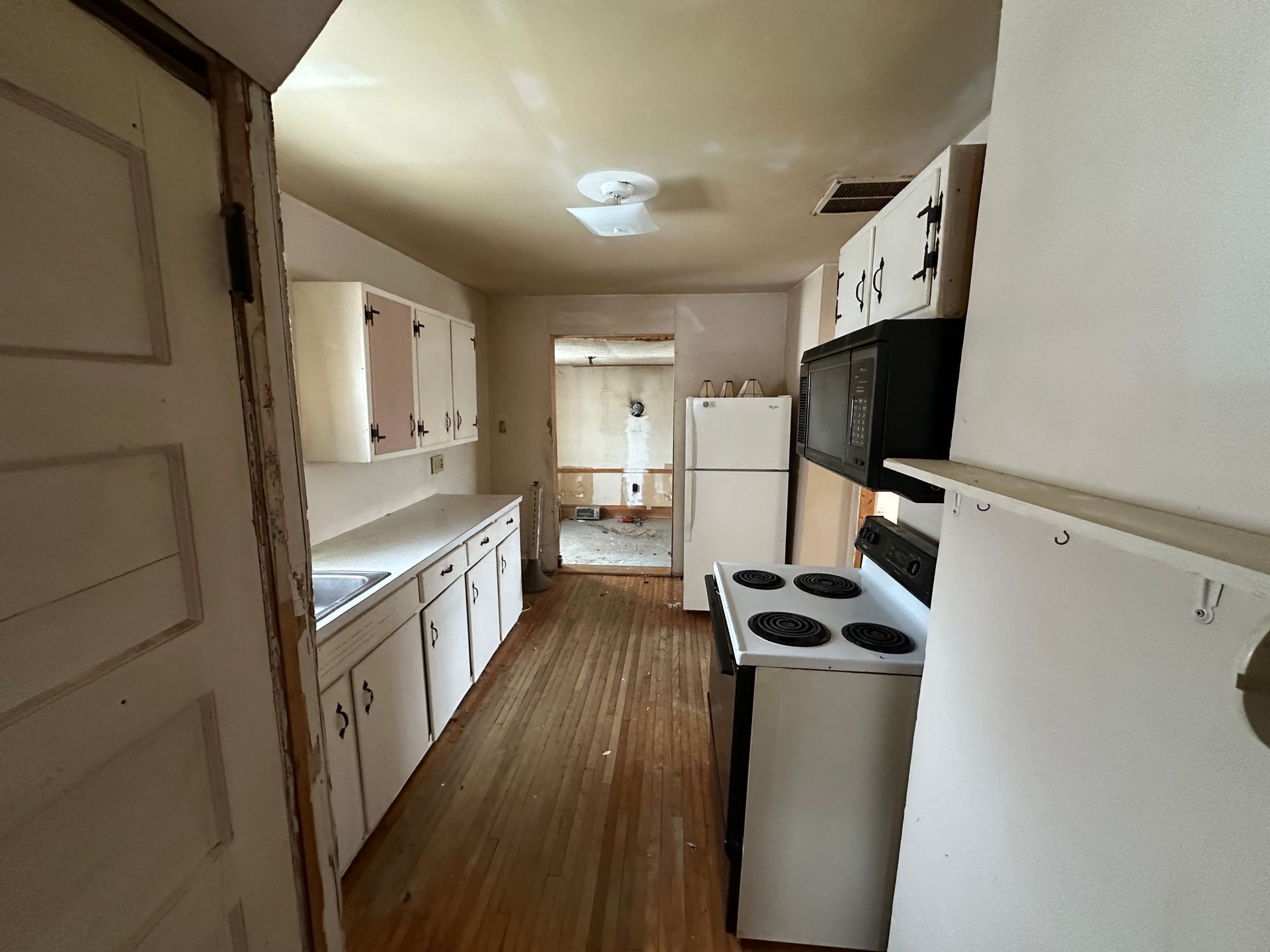 A kitchen with white cabinets, a stove, a refrigerator, and wooden floors, leading into another room.