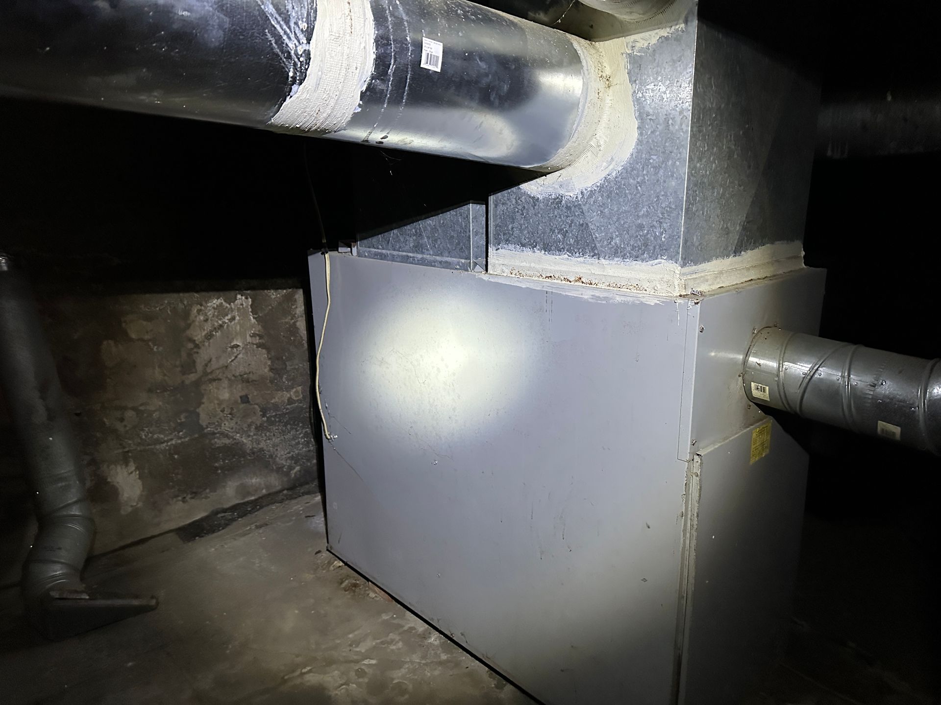 A grey HVAC air handler unit in a dark basement with connected metal ductwork.