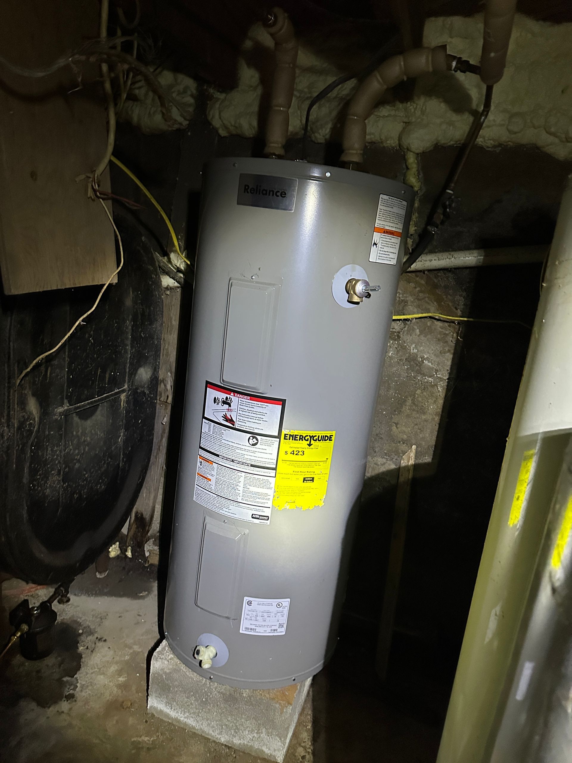 A grey residential electric water heater installed on a concrete block.