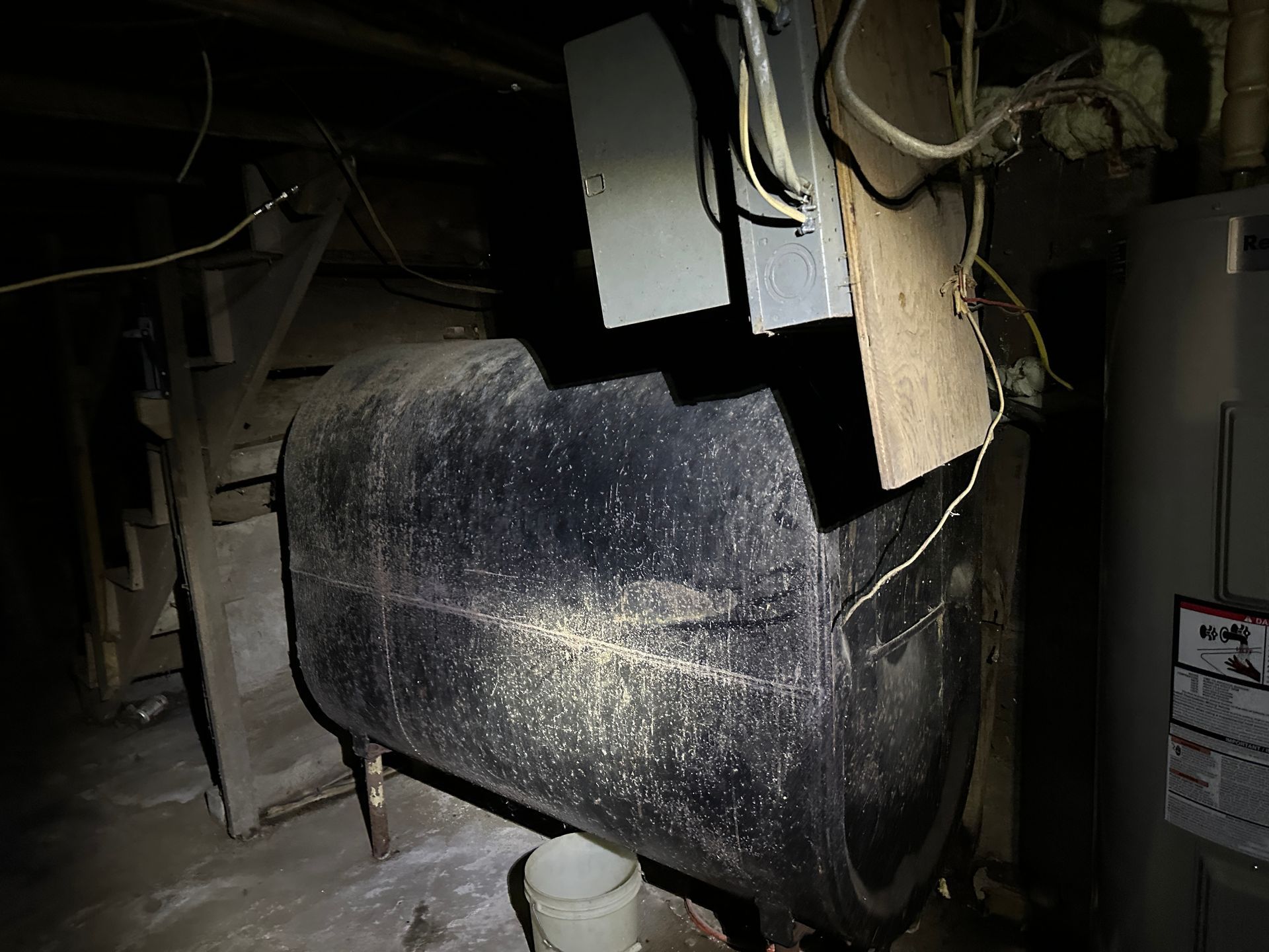 A large, black, horizontal cylindrical tank in a dark basement with electrical equipment mounted on a wooden board above.