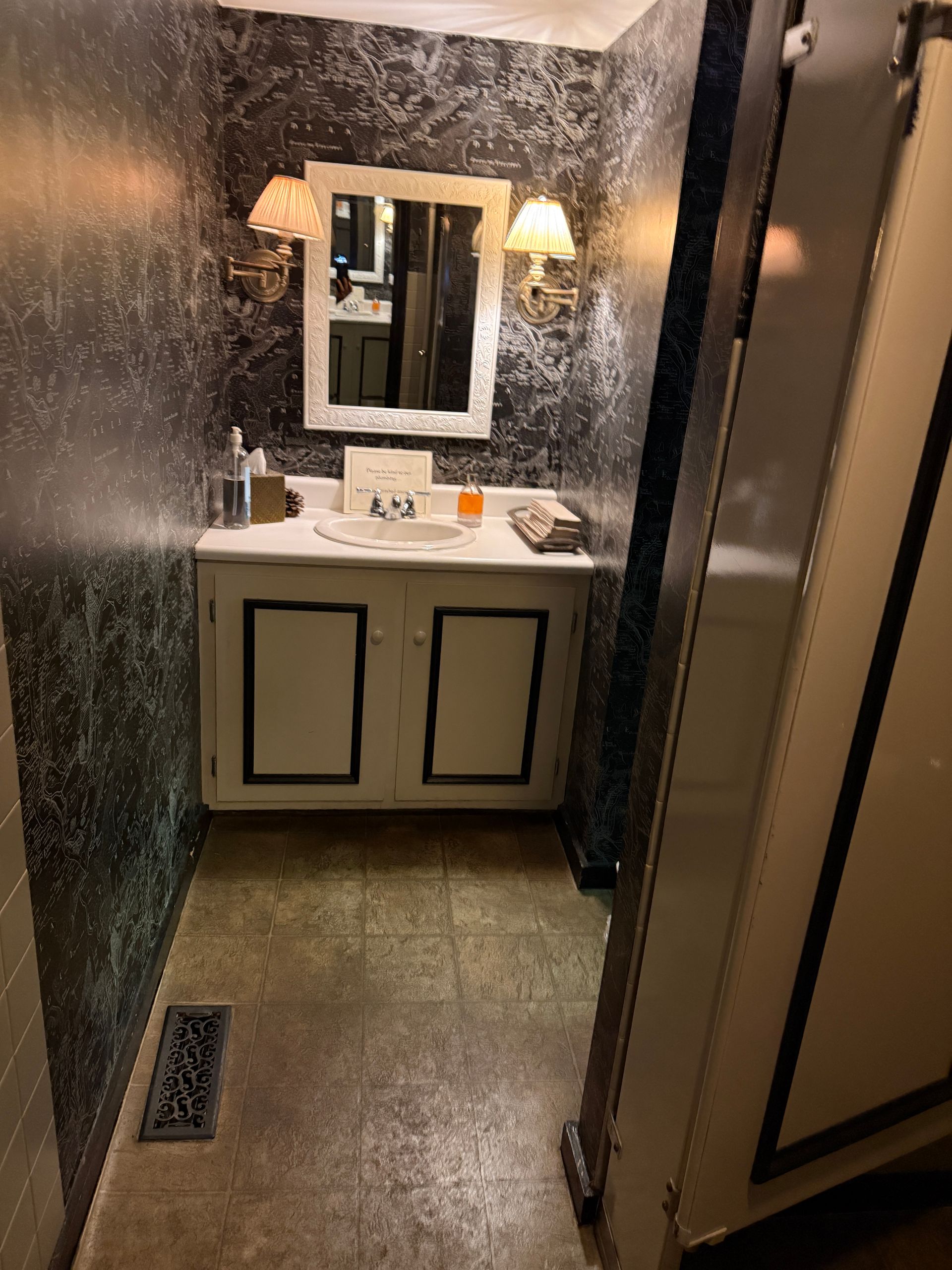 Small restroom with black walls, white vanity and mirror, lit by a wall sconce, seen through a doorway.