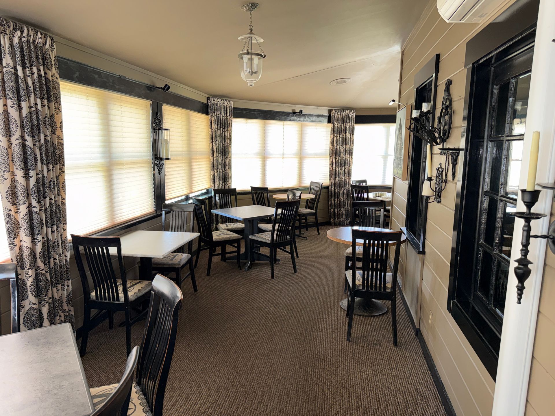 Empty dining room with wicker chairs, tables, and large windows with patterned curtains.