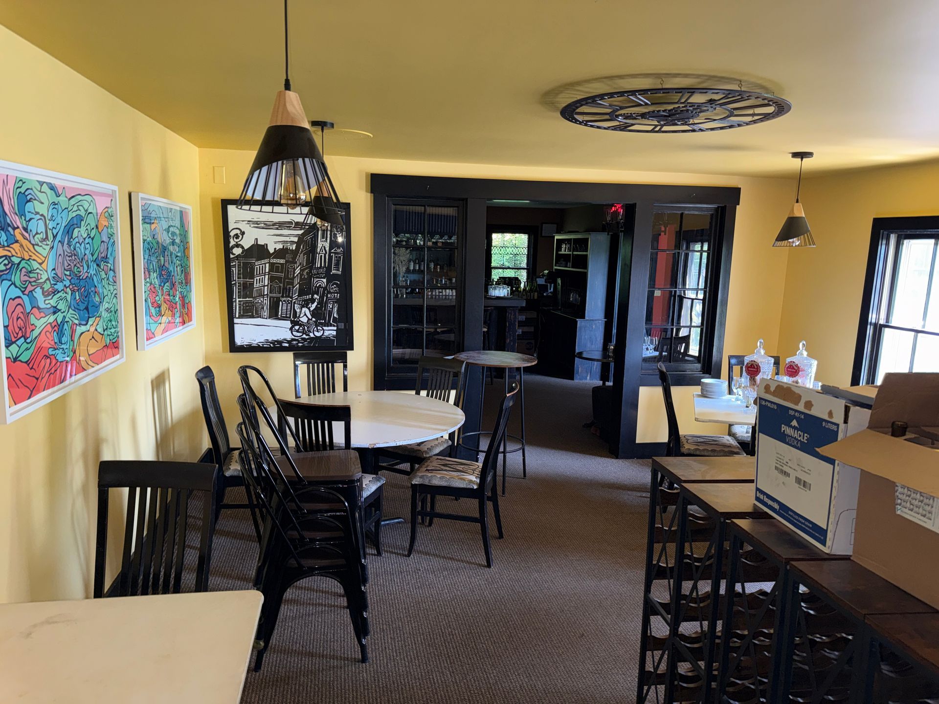 Indoor cafe dining area with black chairs, round tables, warm yellow walls, and colorful artwork.
