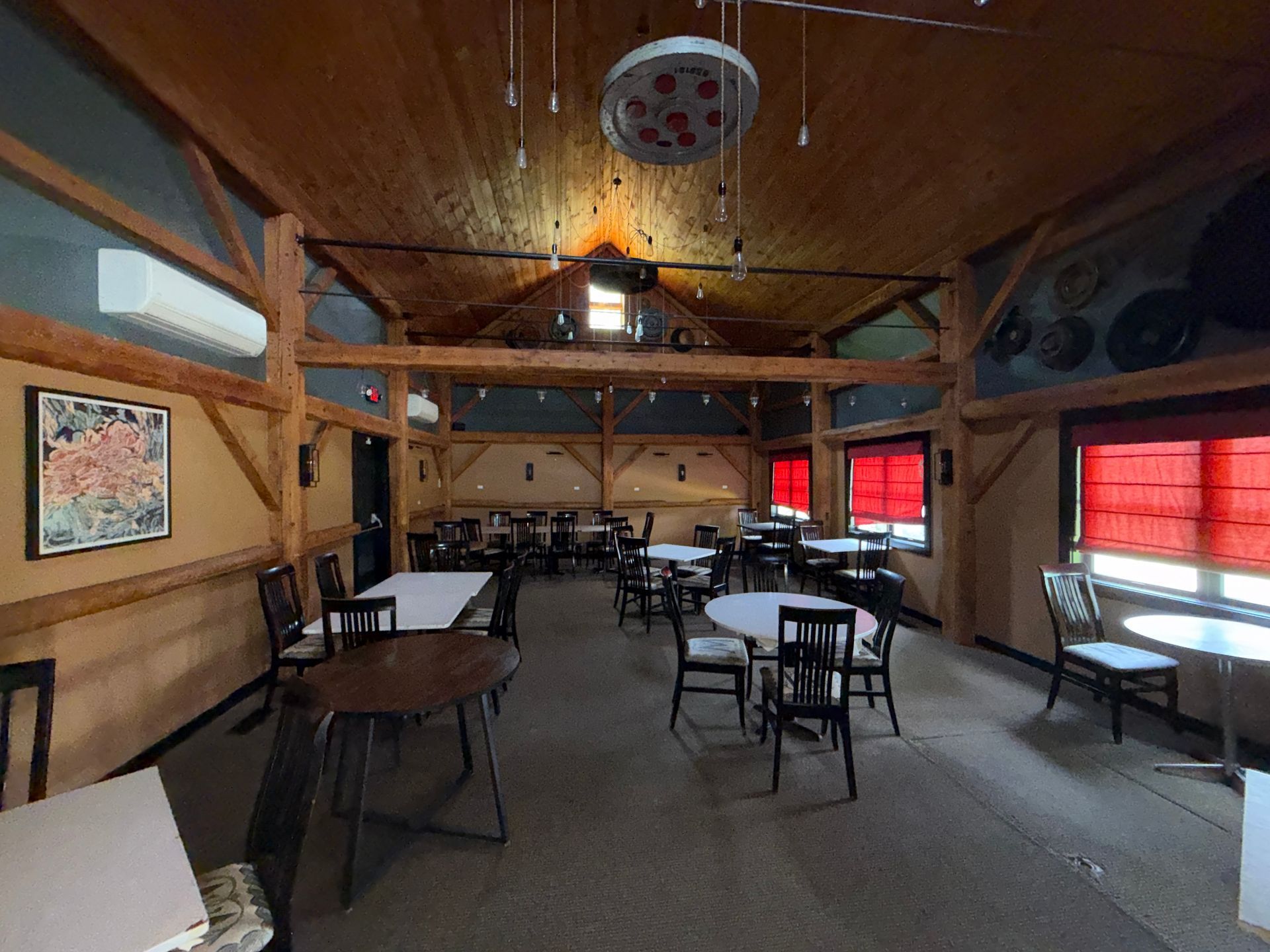 Rustic dining hall with wooden beams, tables, chairs, and red-covered windows