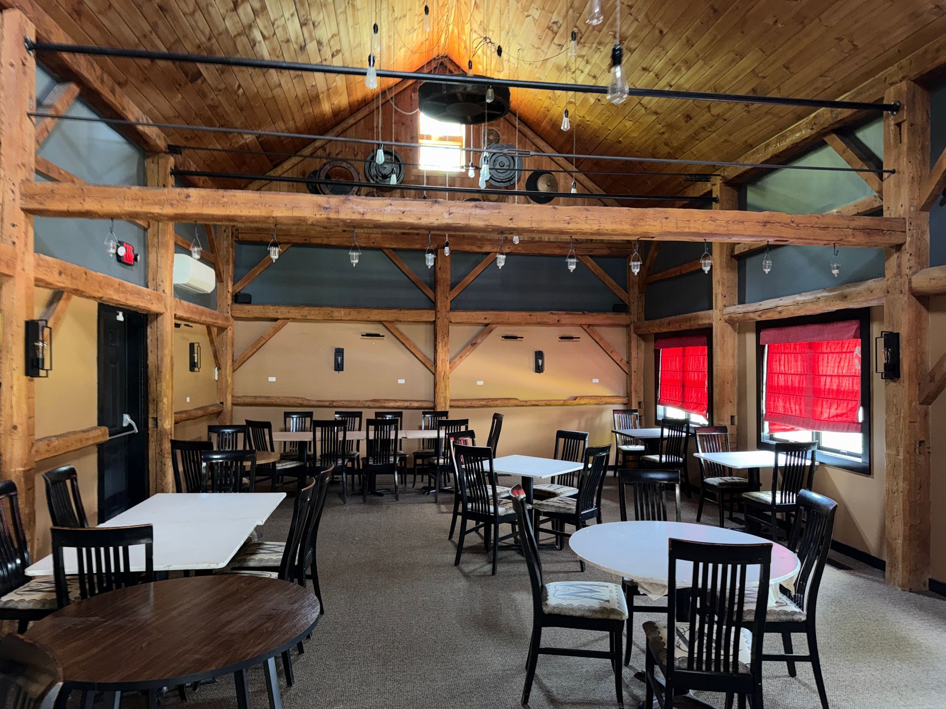 Rustic banquet hall with wooden beams, round tables, and chairs under red-curtained windows.