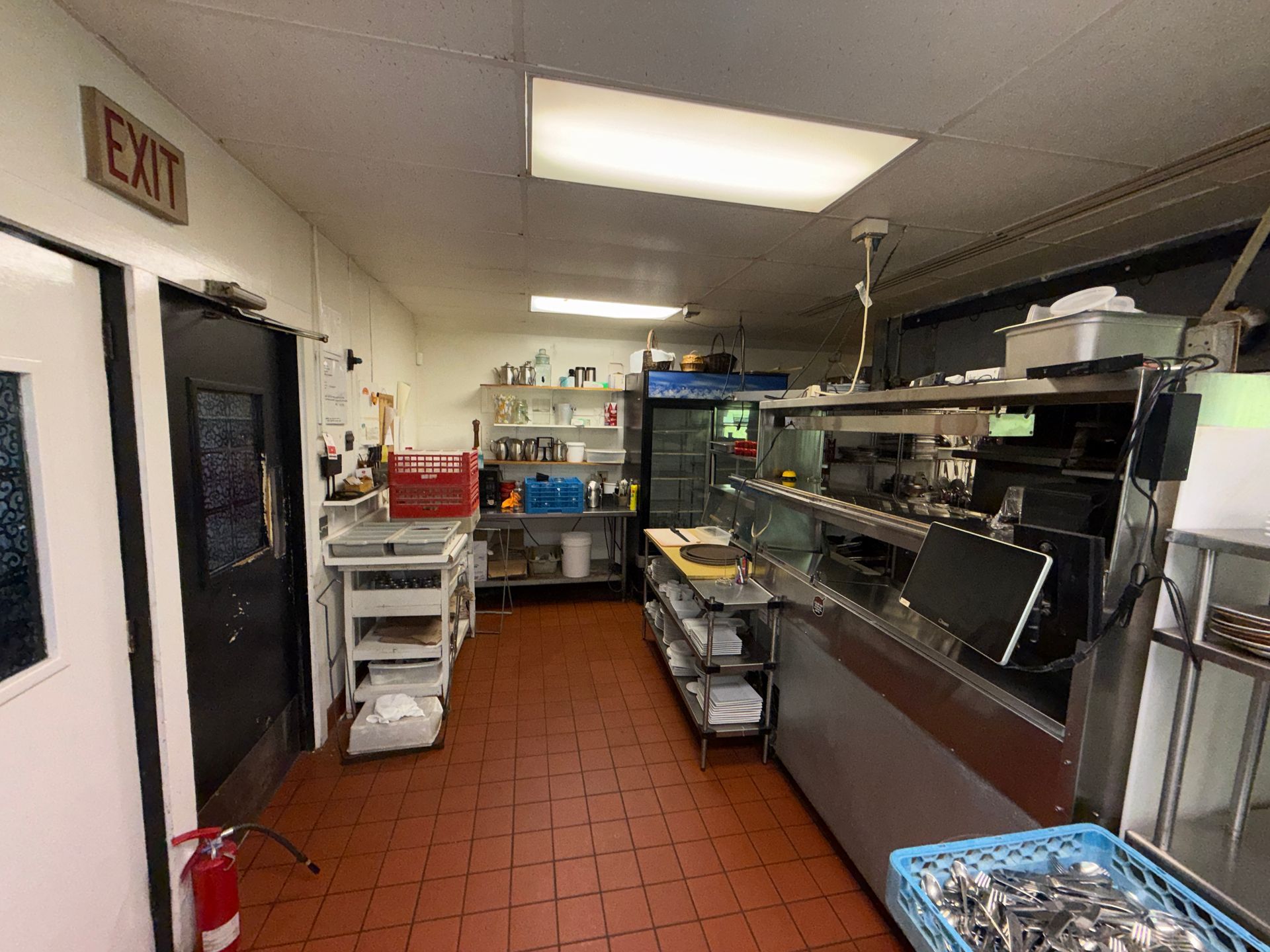 Commercial kitchen with stainless-steel prep tables, shelves, and equipment under bright fluorescent lights.