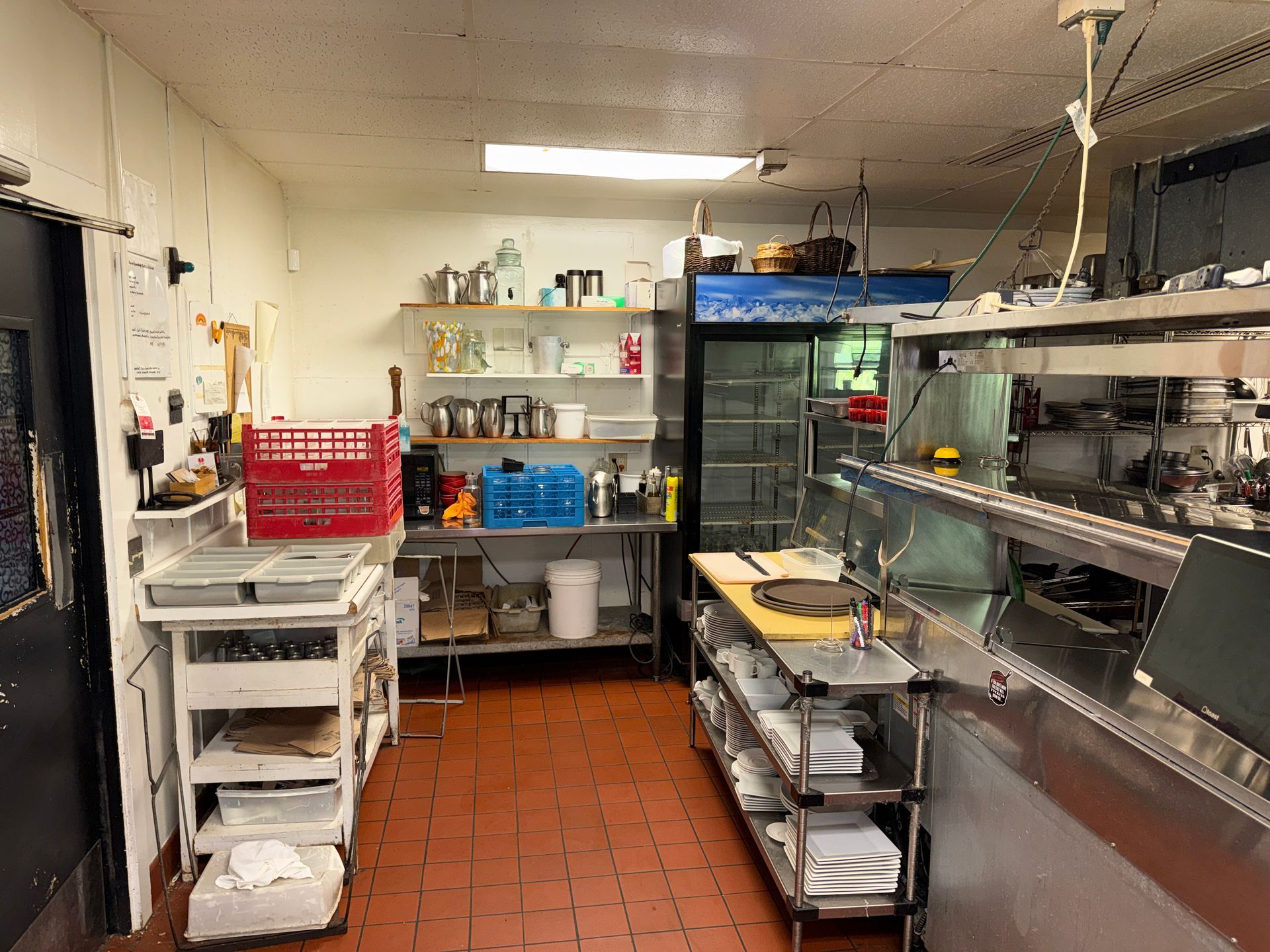 Commercial kitchen with stainless prep tables, shelves, and red tile floor.