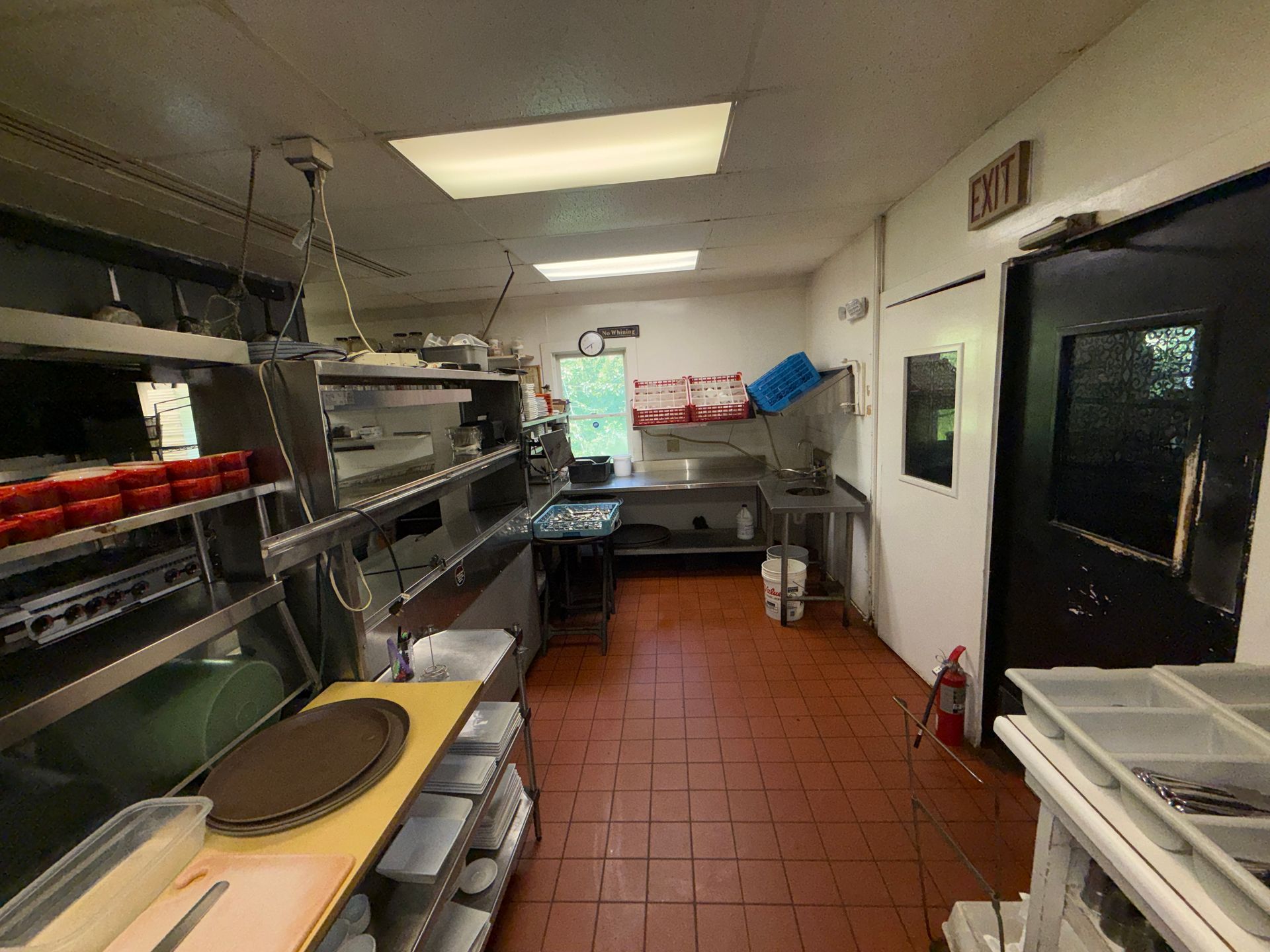 Narrow commercial kitchen with stainless steel counters, shelves, tile floor, and a door at the far end.