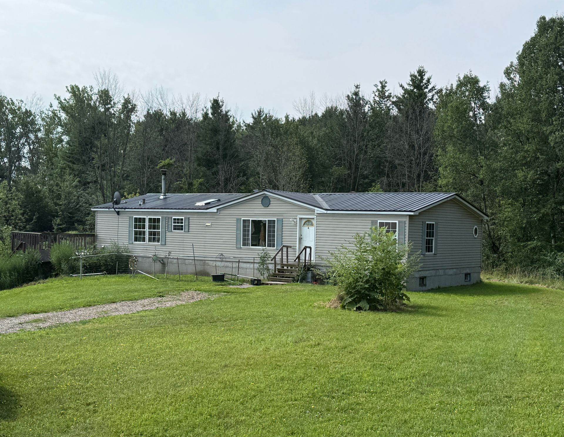 White single-story house with a metal roof on a grassy lot, surrounded by trees under a clear sky