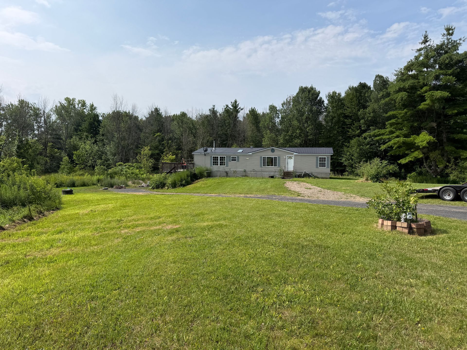 Grassy yard with a long white mobile home, trees, gravel drive, and a blue sky.