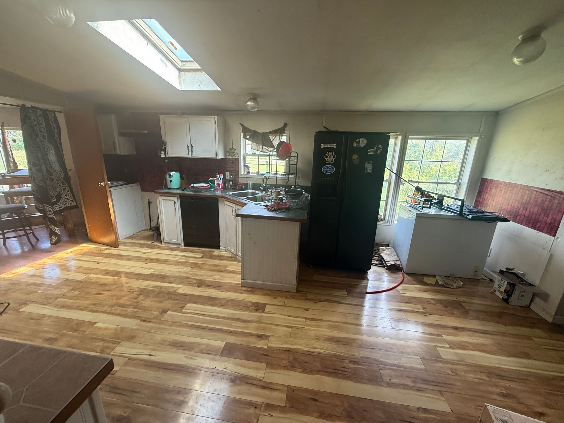 Bright open kitchen and dining area with wood floors, skylight, black fridge, and large windows