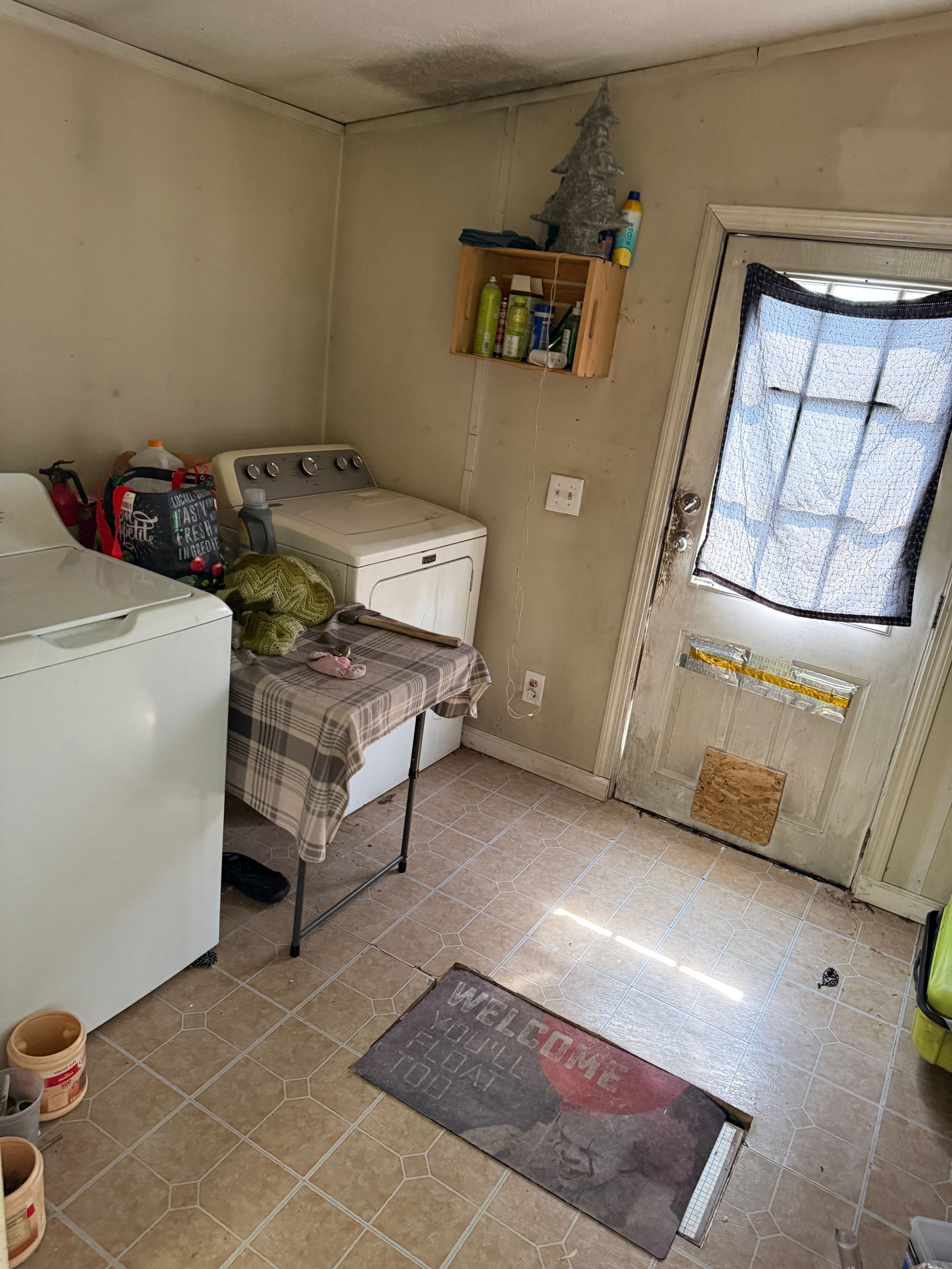 Small beige utility room with freezer, sink, shelves, and a back door with a curtain.