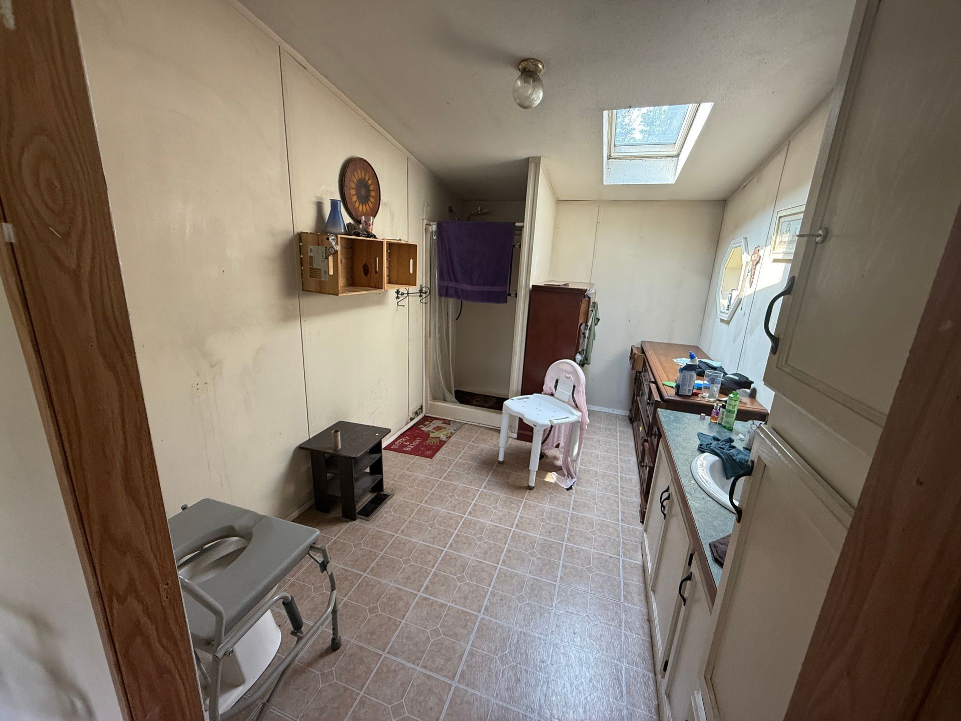 Narrow kitchen corridor with tiled floor, cabinets, chair, shelves, and a skylight overhead.