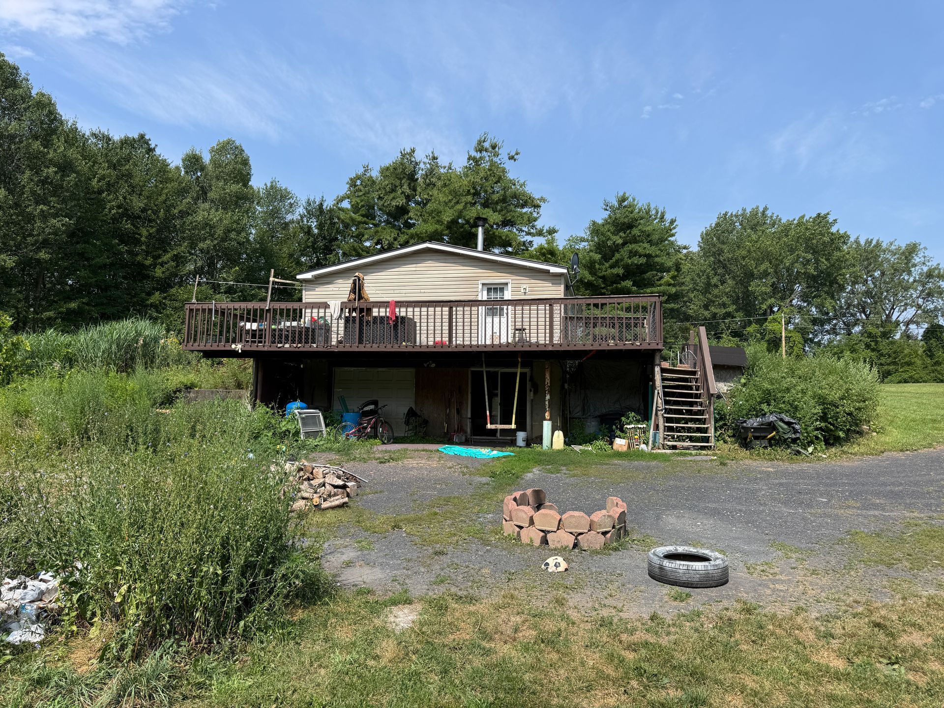 Rustic house with a large deck, surrounded by greenery and a gravel yard under a blue sky