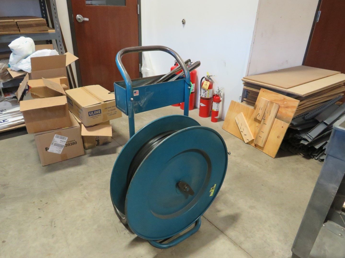 Blue strapping dispenser with a handle, next to boxes, fire extinguishers, and other items in a storage area.