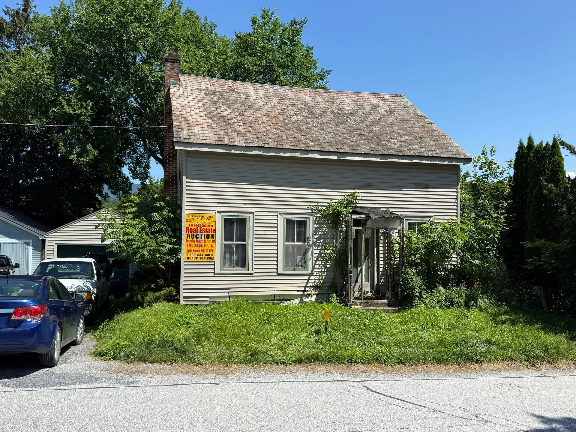 Small white house with a weathered roof and overgrown yard beside parked cars on a sunny street.