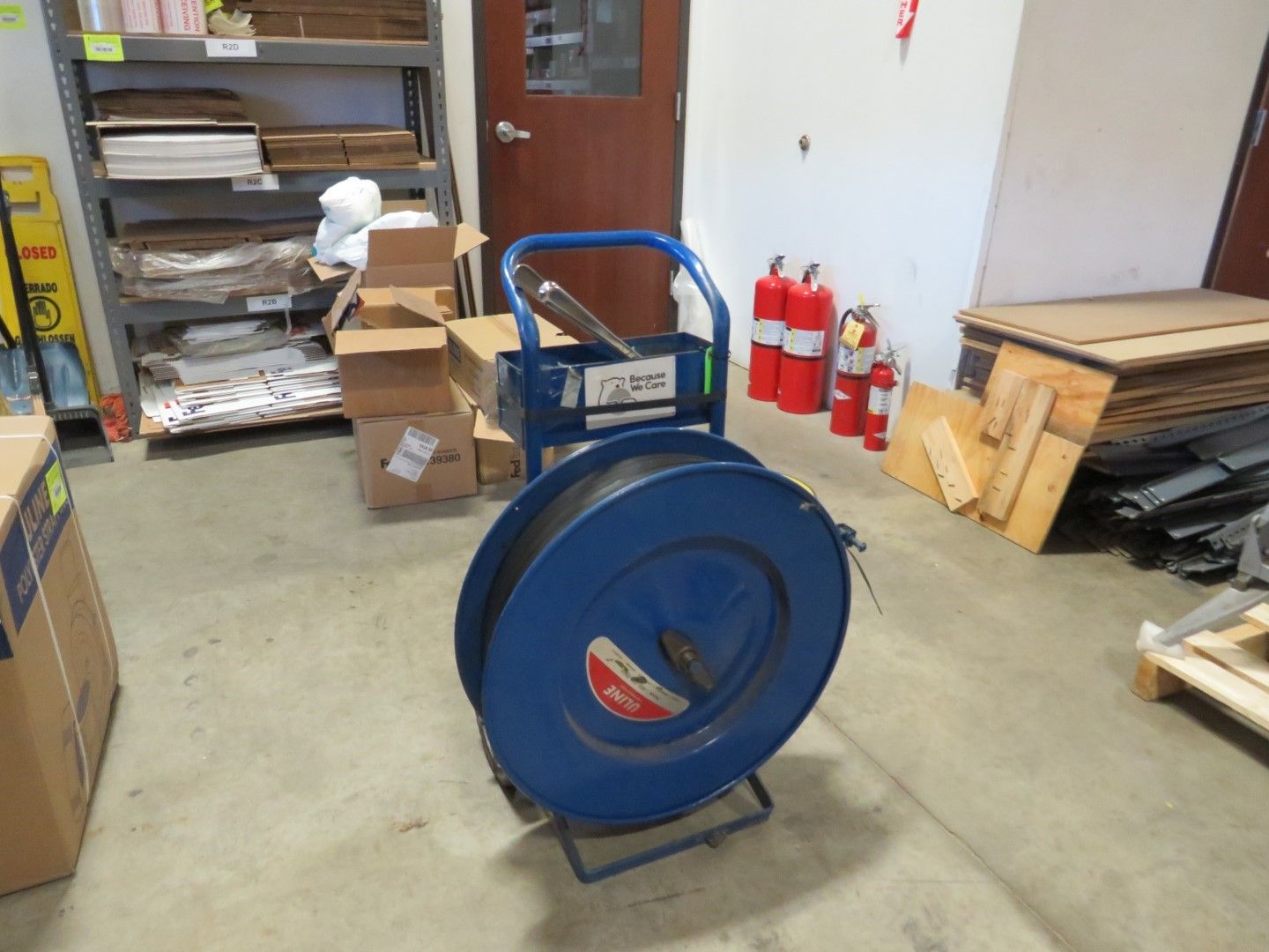 Blue strapping dispenser in a warehouse setting, with boxes, fire extinguishers, and shelving visible.