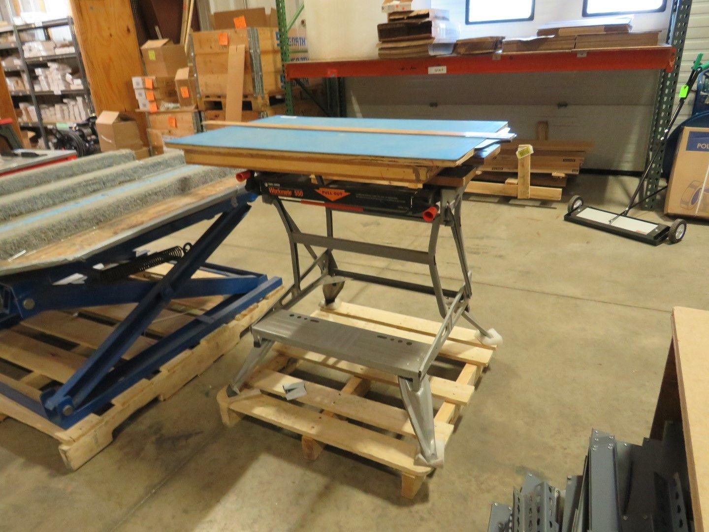 Black and silver workbench on a wooden pallet in a warehouse. Blue surface, various materials in the background.