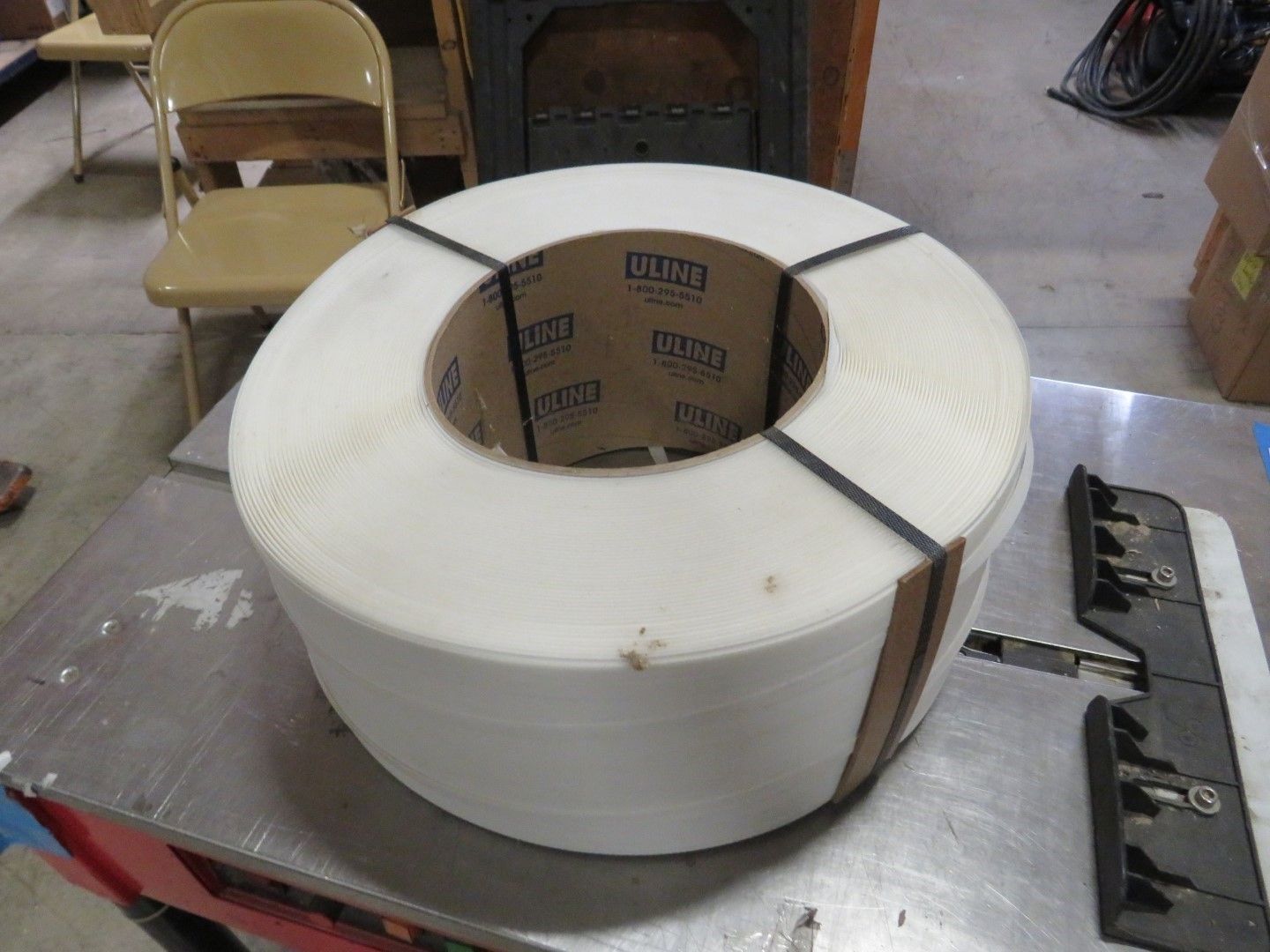 Coil of white plastic strapping on a table, secured with black bands, with a chair in the background.