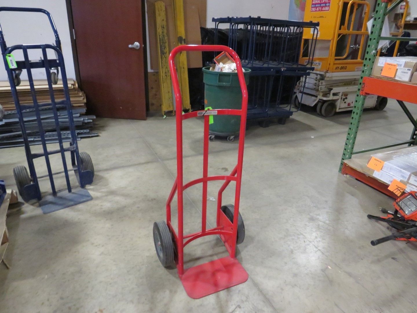 Red hand truck with black wheels, on a concrete floor in a warehouse.