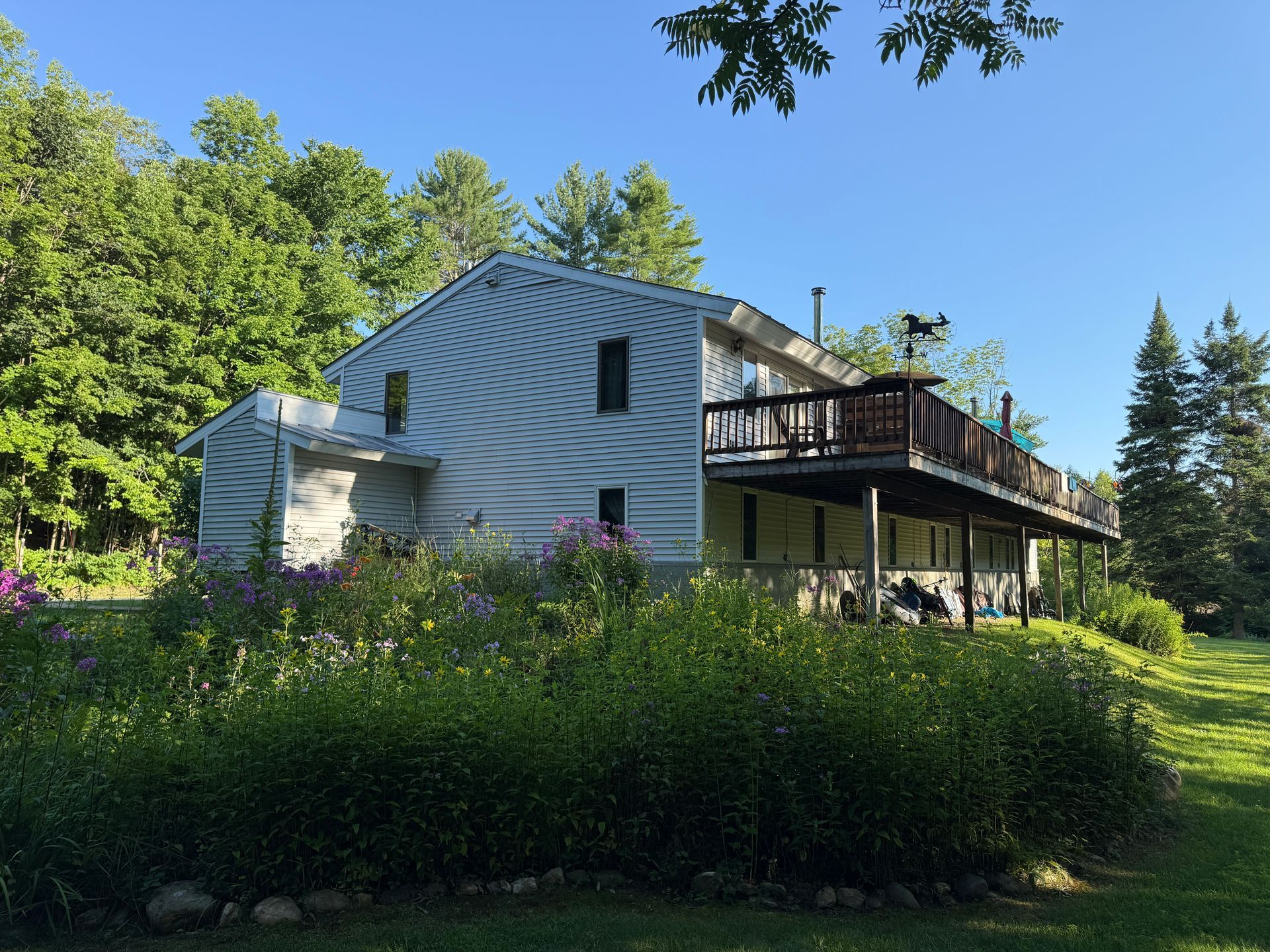 White house with a wooden deck on a sunny, leafy hillside under a clear blue sky