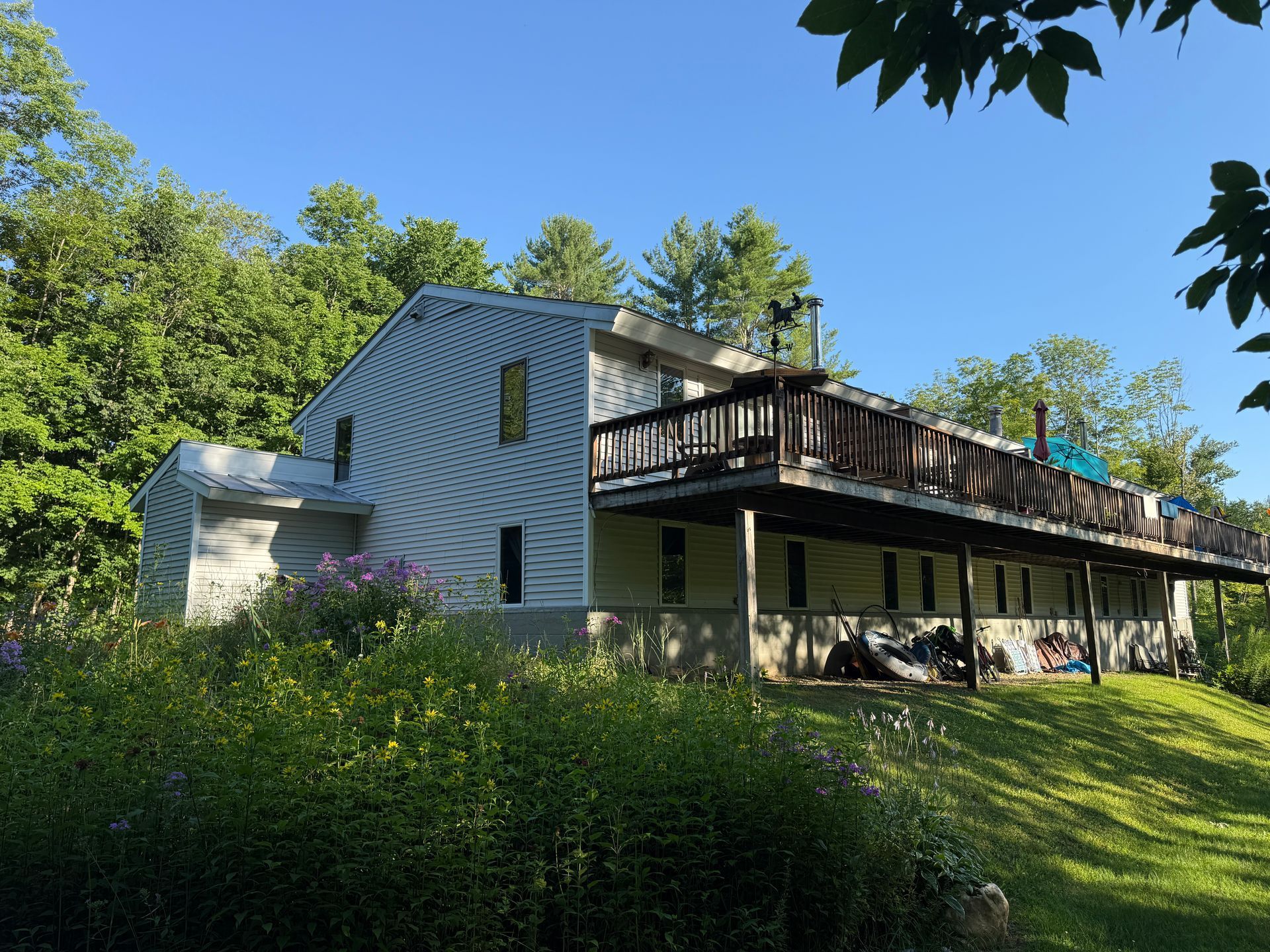 White house with a dark wooden deck on a grassy, tree-lined hillside under a blue sky