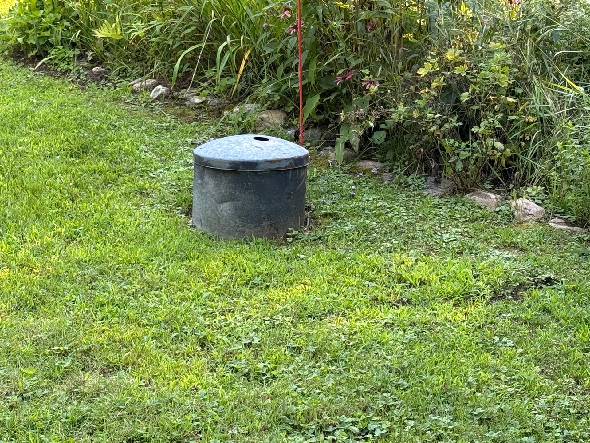 A small black cylindrical utility cover in a grassy yard beside bushes.