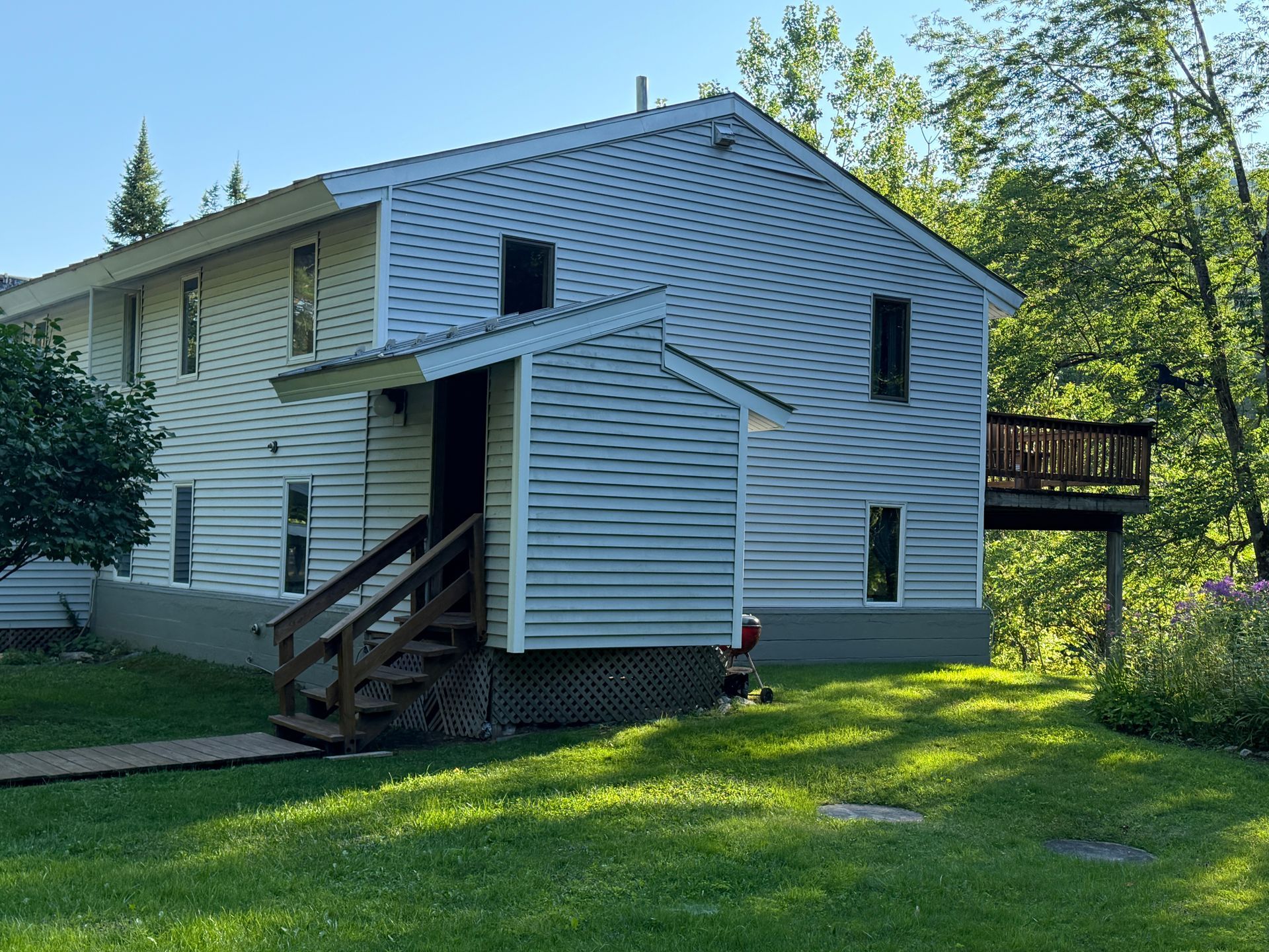 White house exterior with wooden stairs, side deck, and a person sitting on the lawn in a sunny yard