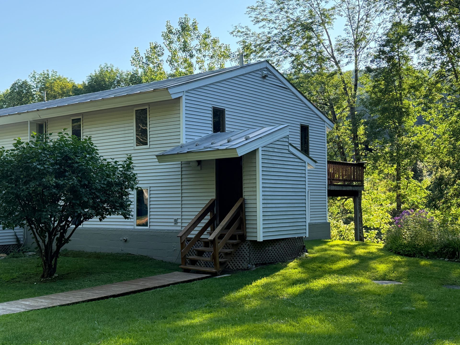 White two-story house with gray siding, small front porch, and green lawn surrounded by trees.