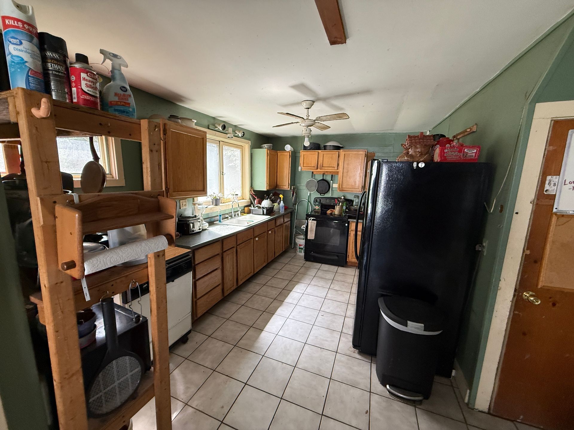 Narrow kitchen with wooden cabinets, tiled floor, black refrigerator, and daylight from windows.