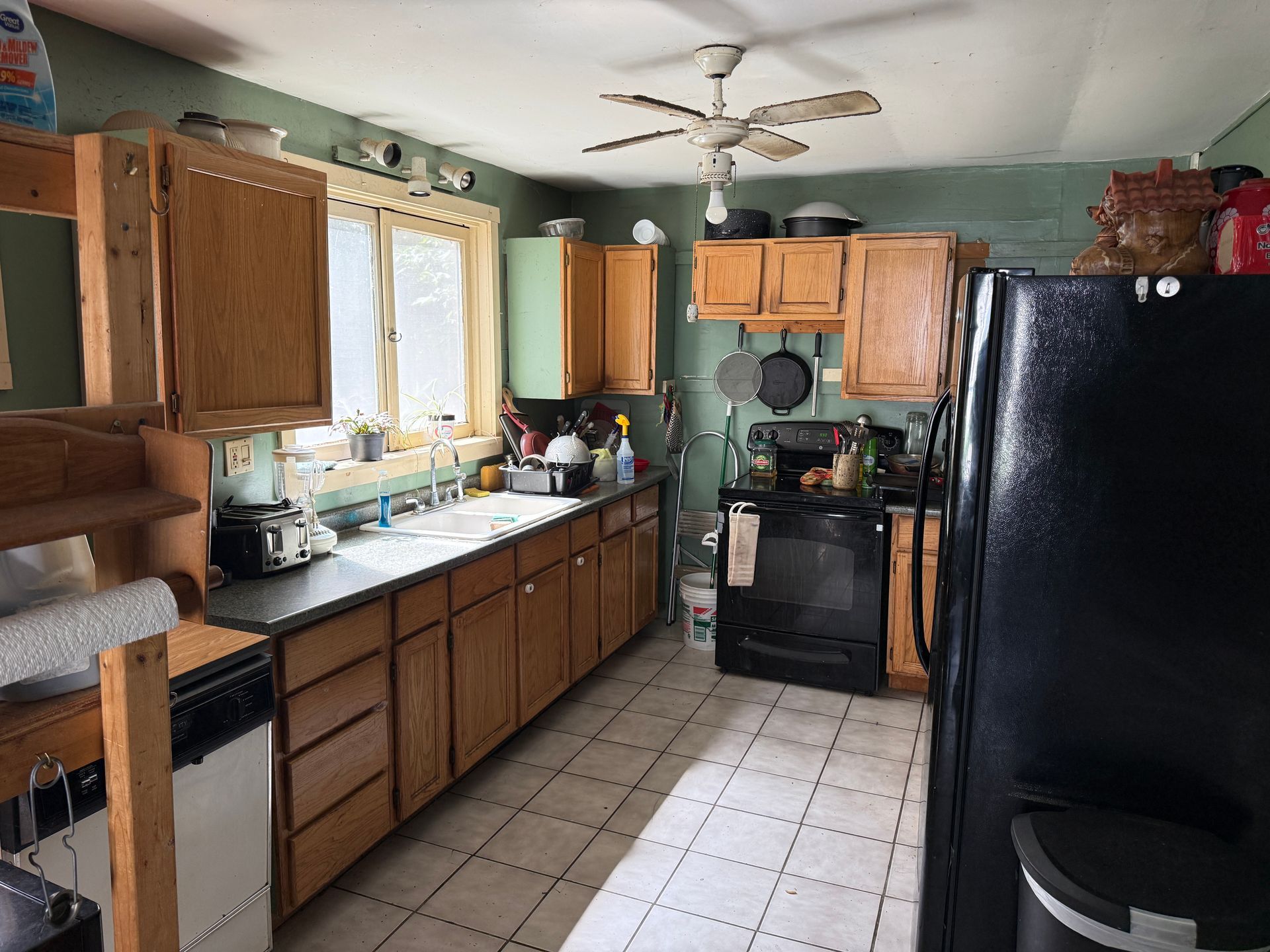Small kitchen with wooden cabinets, black stove and fridge, white tile floor, and daylight from a window.