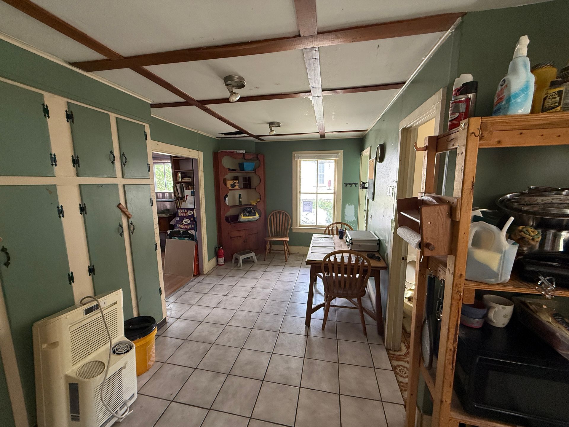 Narrow kitchen or hallway with tiled floor, green walls, wooden shelves, and a small table near a bright window.