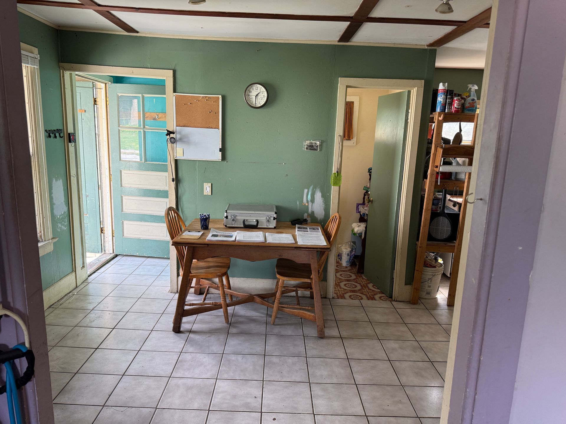 Small kitchen with green walls, tiled floor, wooden table, chairs, and open doors to adjoining rooms.
