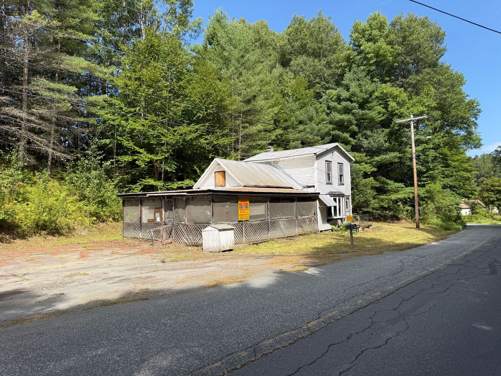 Small white house with porch by a roadside, surrounded by trees and gravel driveway