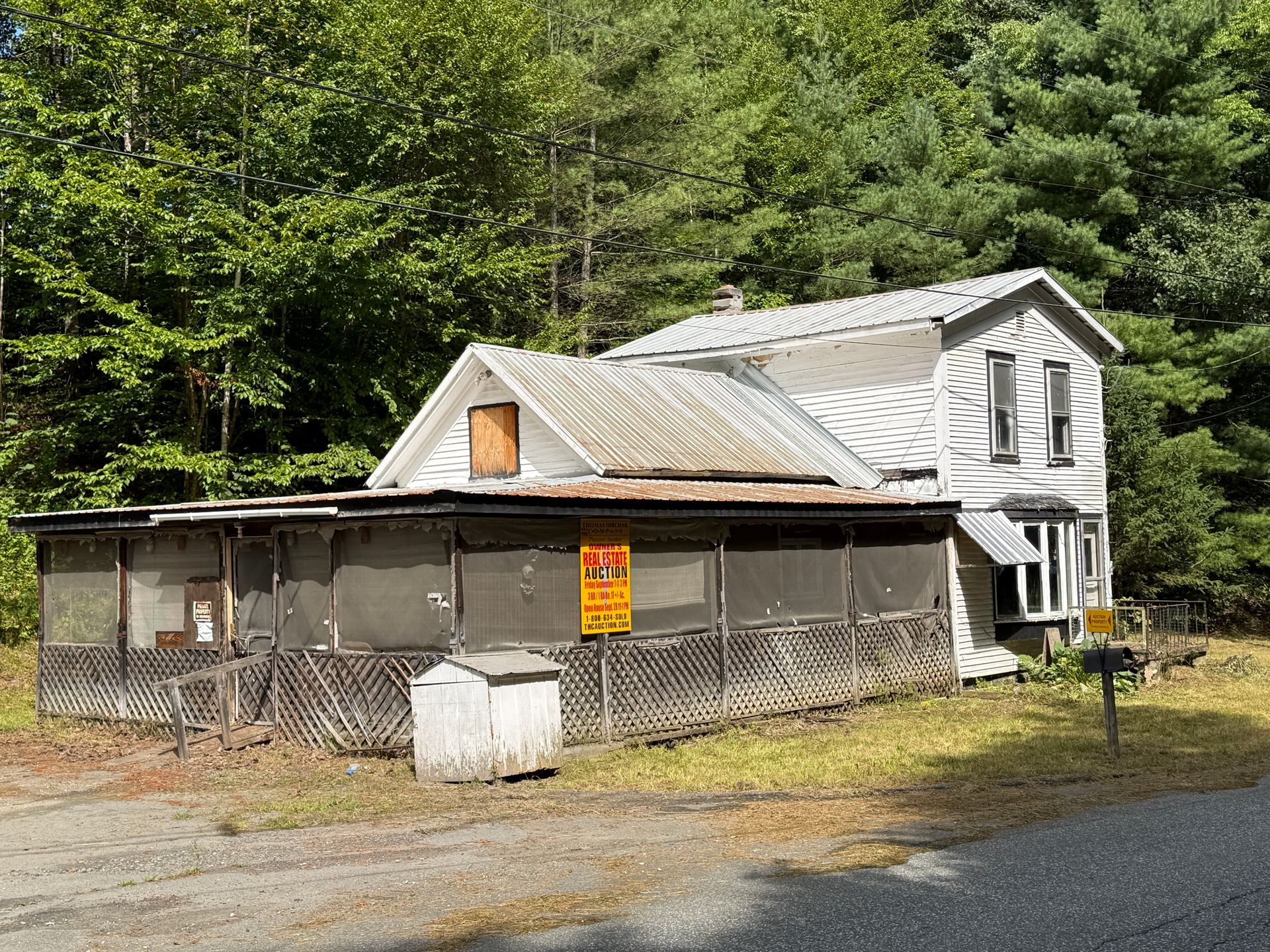 white house with a fenced porch beside a gravel road, surrounded by dense green trees.