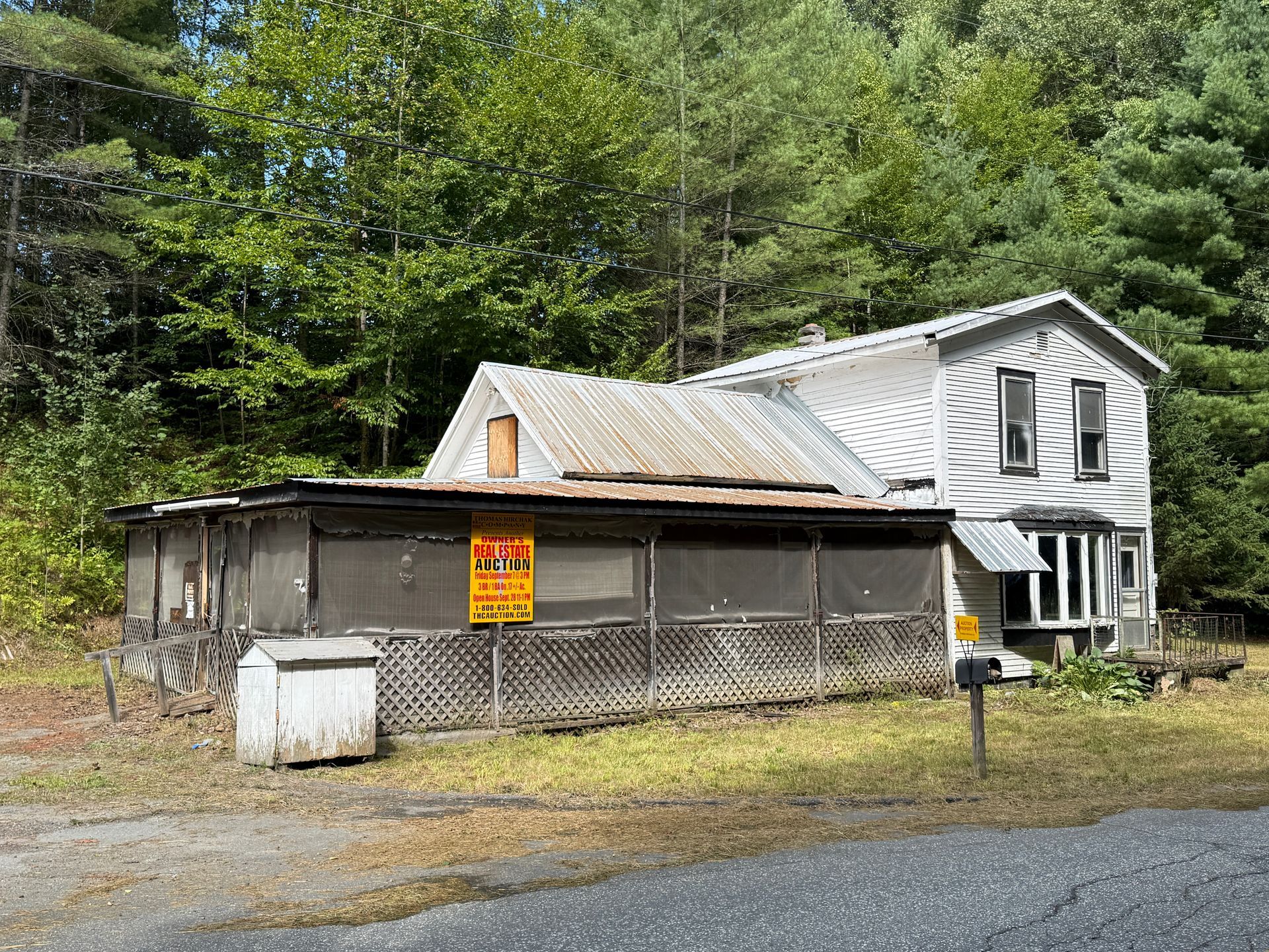 white house with attached shed and mesh fence, set beside a gravel lot and wooded hillside.