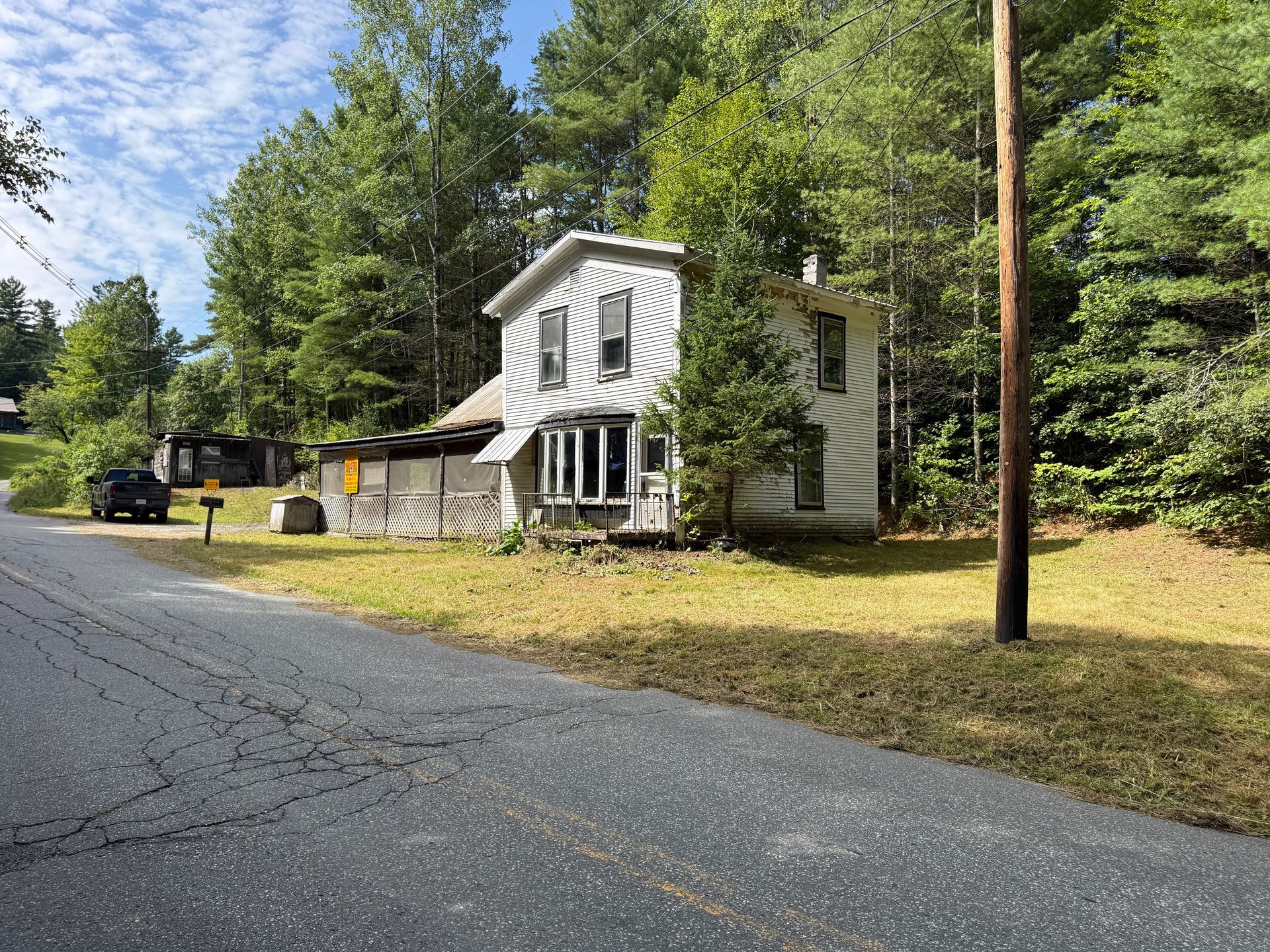 White two-story house beside a quiet road, surrounded by trees and a grassy yard.