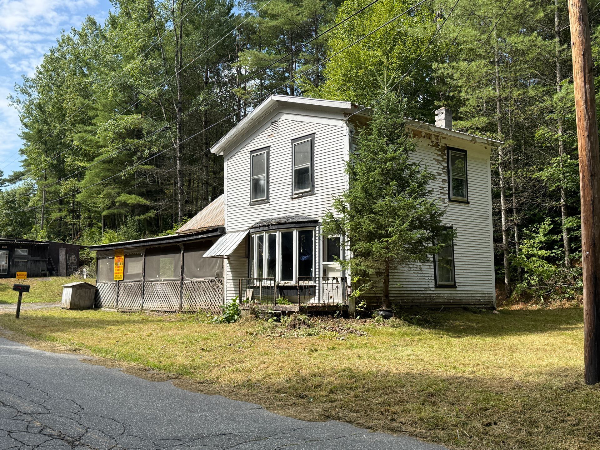 Old white house with a porch beside a road, surrounded by trees in a wooded yard