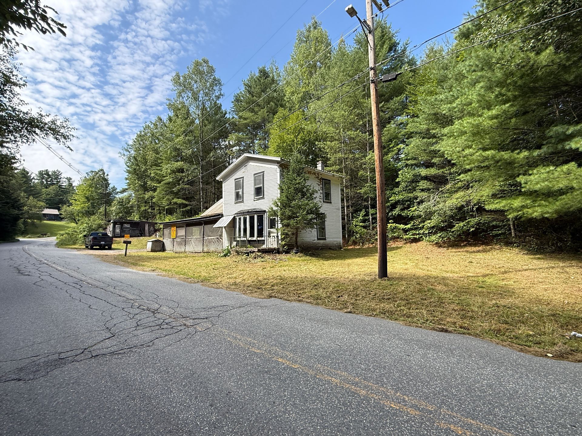 Quiet rural road beside a small white house with trees and utility poles under a blue sky