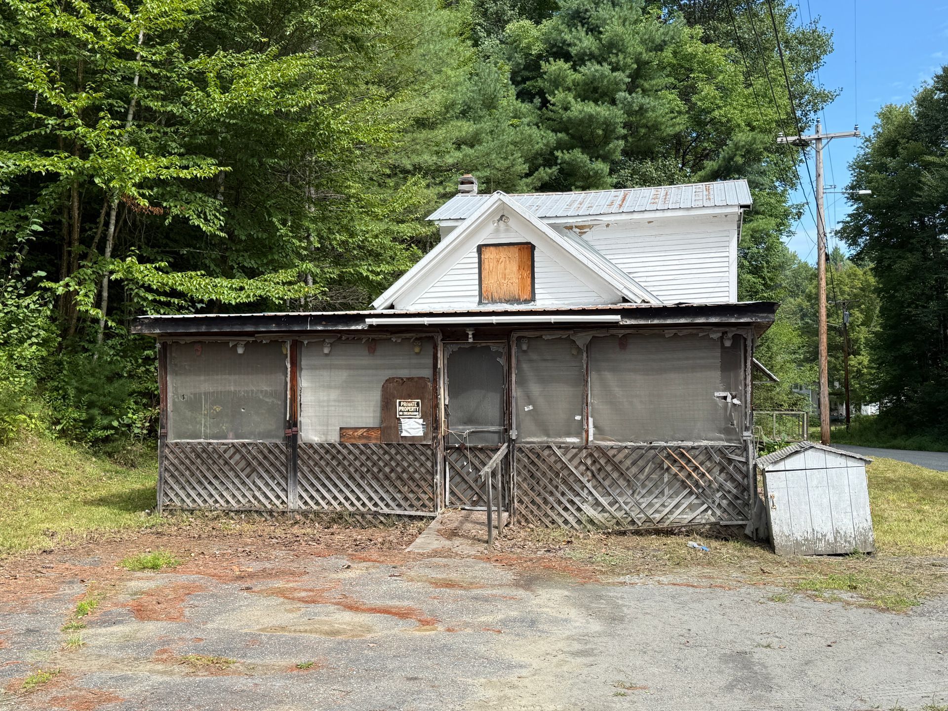Small roadside house with a porch, set against trees and gravel.