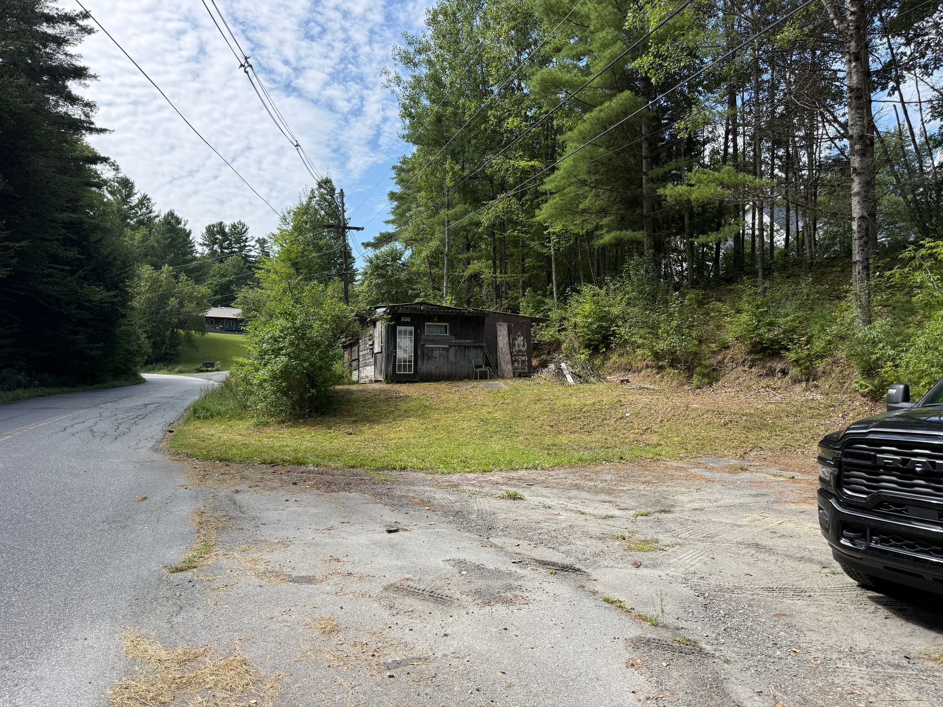 Gravel road curving past trees and a small shed, with the front of a black SUV at right.