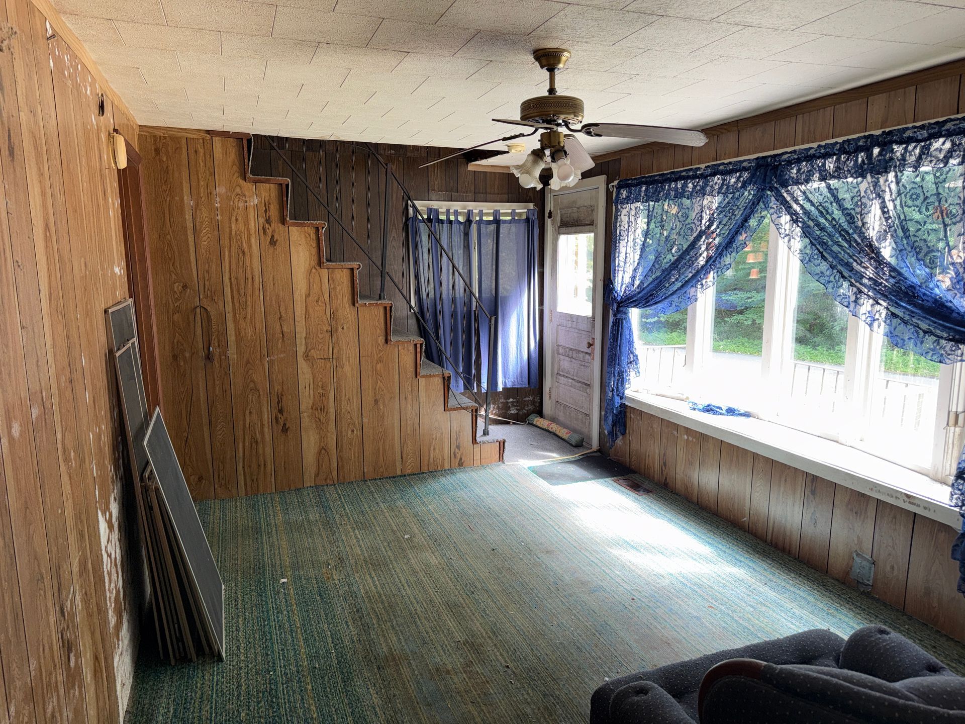 Empty living room with green carpet, wood-paneled walls, large windows, and a ceiling fan
