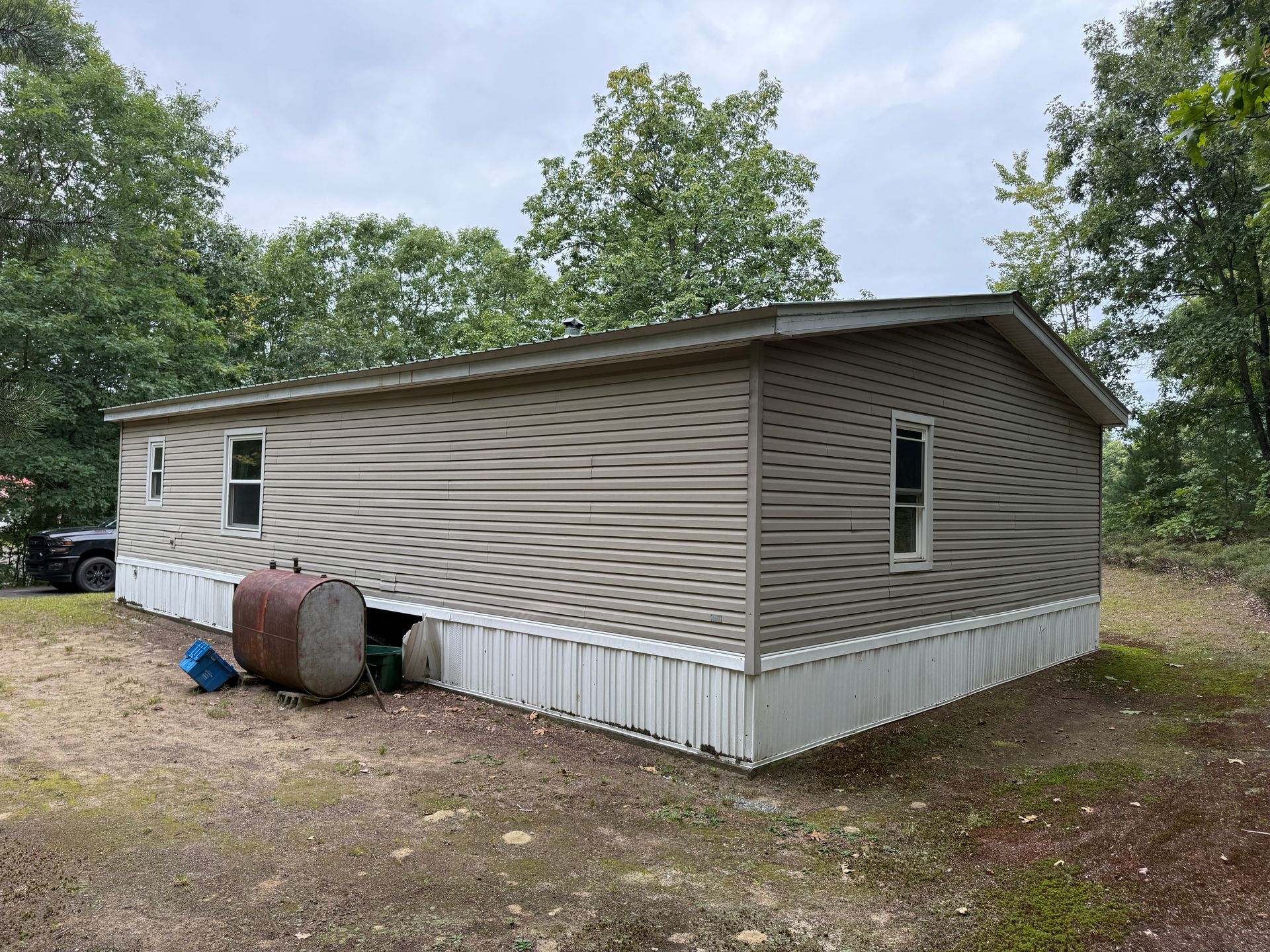 Tan mobile home with beige siding and white skirting in a wooded lot under a cloudy sky