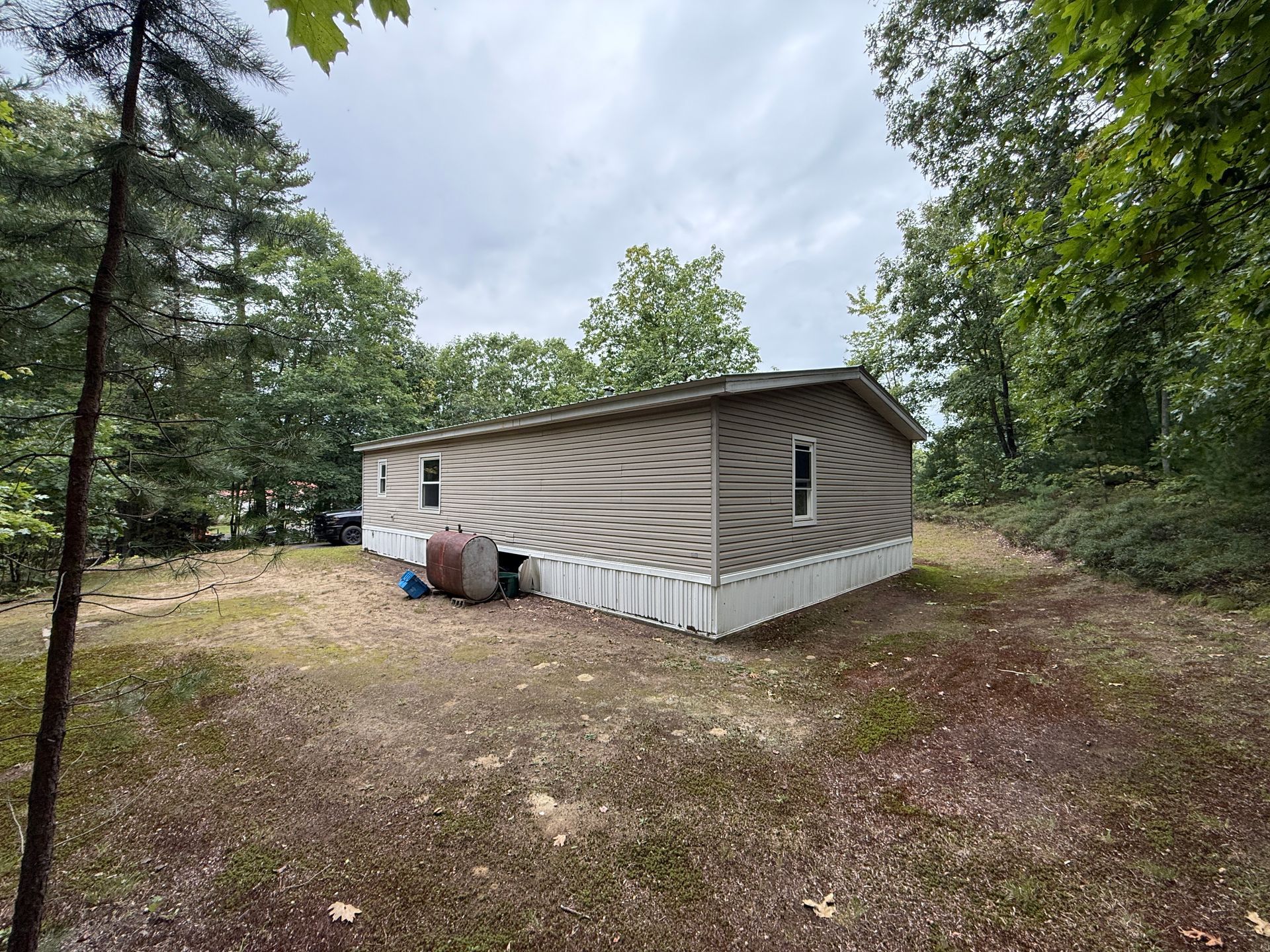 Single-story beige mobile home in a wooded clearing with a white skirting and a small porch.